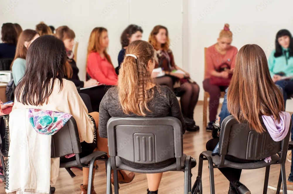Group of women sitting in a circle during a meeting or workshop in a room with plain white walls.