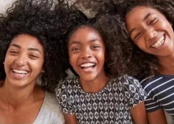 Three young women smiling and laughing together, with curly hair, in casual clothing.