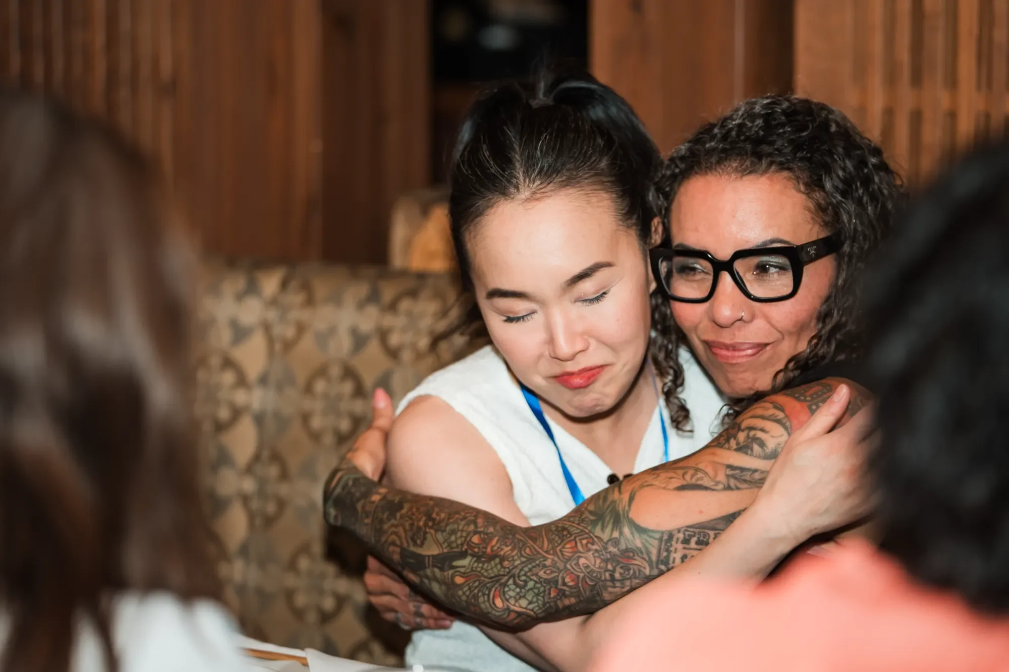 Two women sharing a warm embrace, smiling, in a cozy indoor setting with wood-paneled walls, one woman with glasses and tattoos, the other with closed eyes and a content expression.