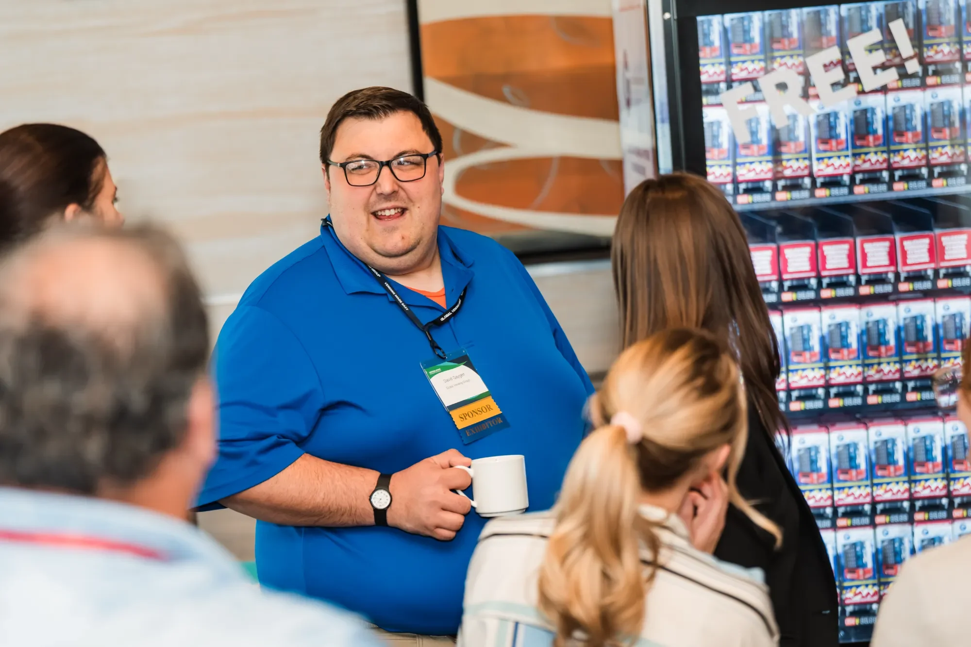 A man with glasses, wearing a blue shirt, holding a white mug, speaking to a group of people at a meeting or conference. There is a large vending machine in the background with the word 'FREE' on it.