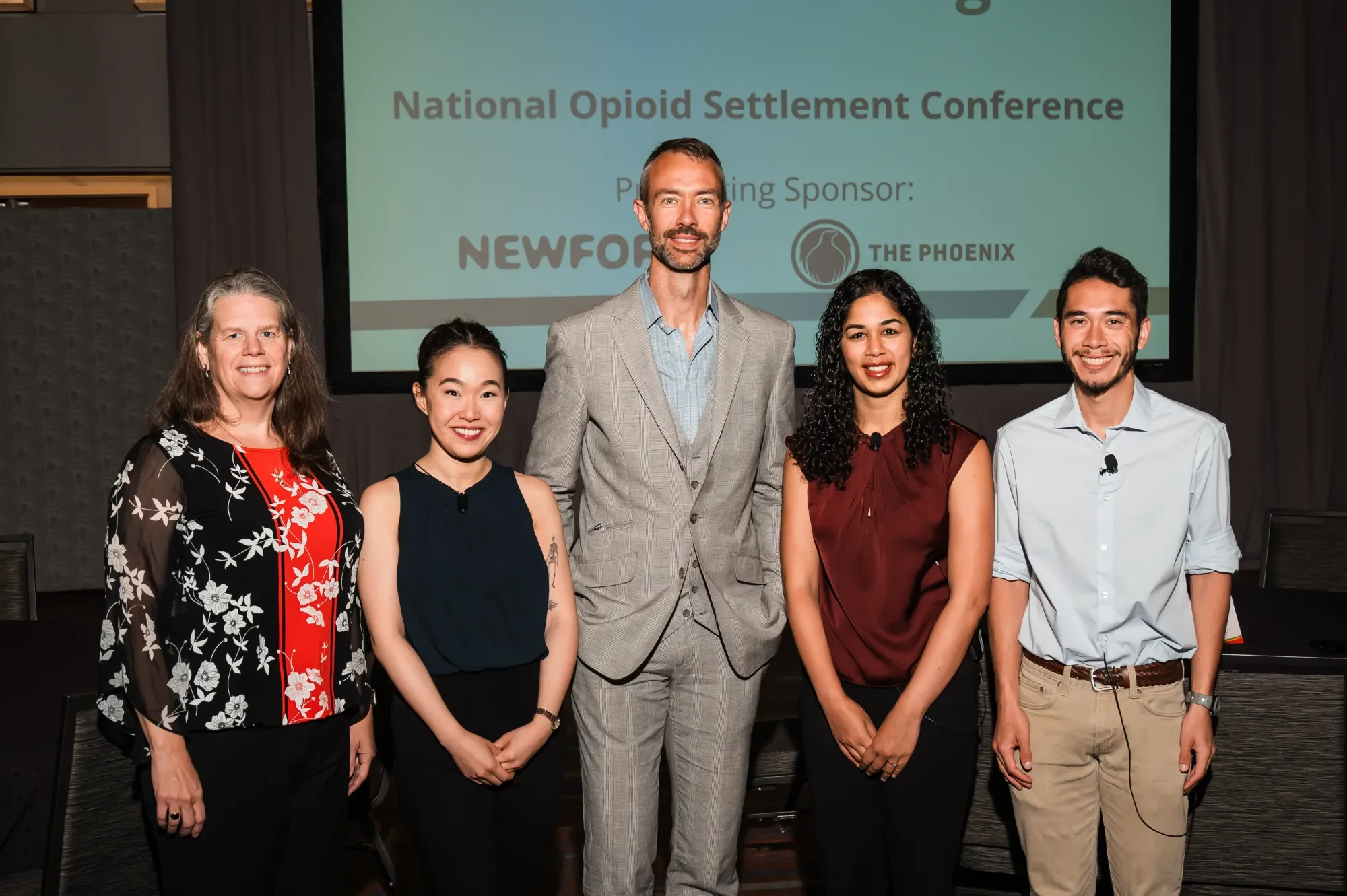 NOSC team standing on stage at a conference with a large screen behind them that reads 'National Opioid Settlement Conference' and lists a sponsor.