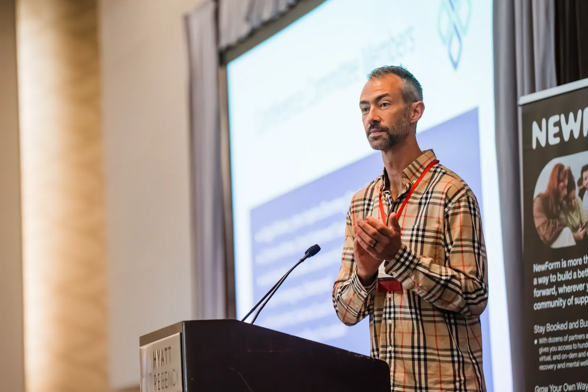 A man with a beard, wearing a plaid shirt, is giving a presentation at a conference or seminar. He is standing behind a podium with a microphone, in front of a large screen displaying a blue and white slide. There is a poster with the word 'NEW' on a