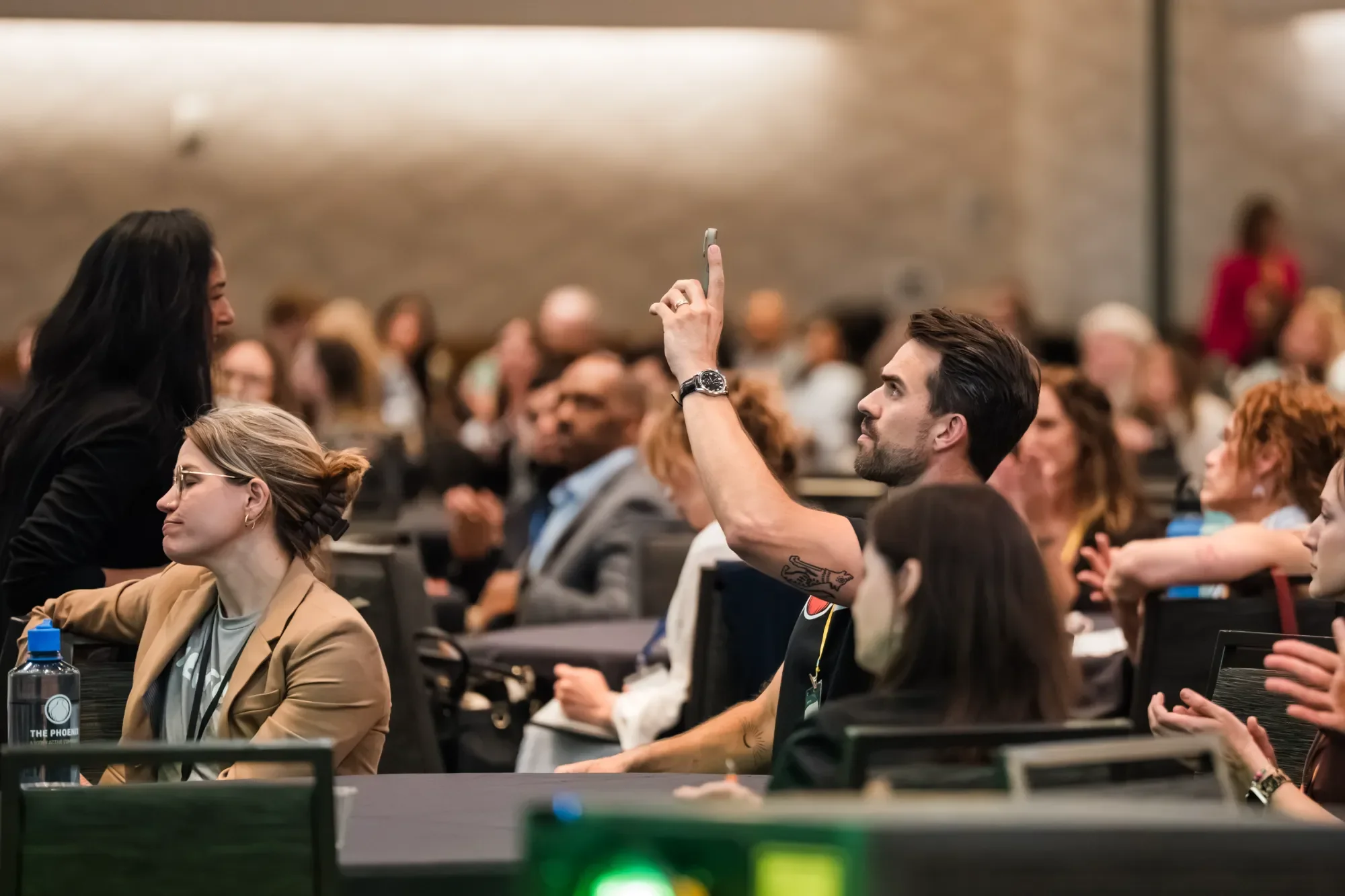 A man with a beard and tattoos on his arm, wearing a watch, is holding up a smartphone to take a photo or video of a woman sitting at a conference or seminar. The woman has long dark hair and is smiling. The background shows many other attendees seat