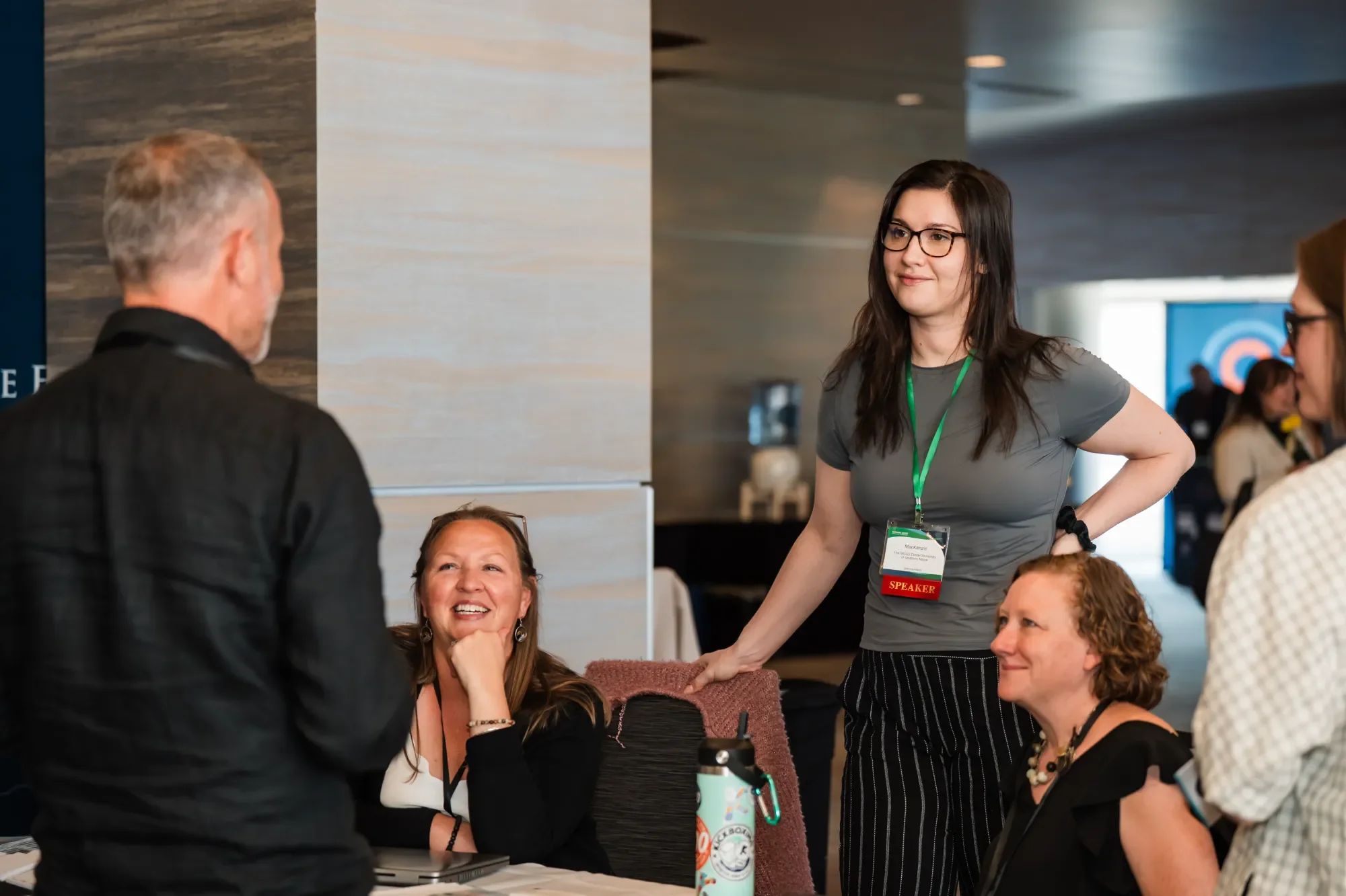 A group of women sitting at a table in a conference, smiling and engaging in conversation. A young woman with glasses and a conference badge labeled 'SPEAKER' stands next to the table, talking to the women.