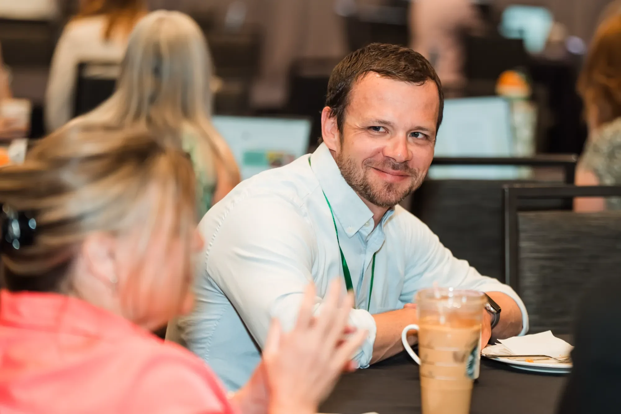 A man in a white shirt smiling and engaged in conversation at a table with coffee drinks in front of him. The scene appears to be in a busy office or cafeteria setting with people working on computers in the background.