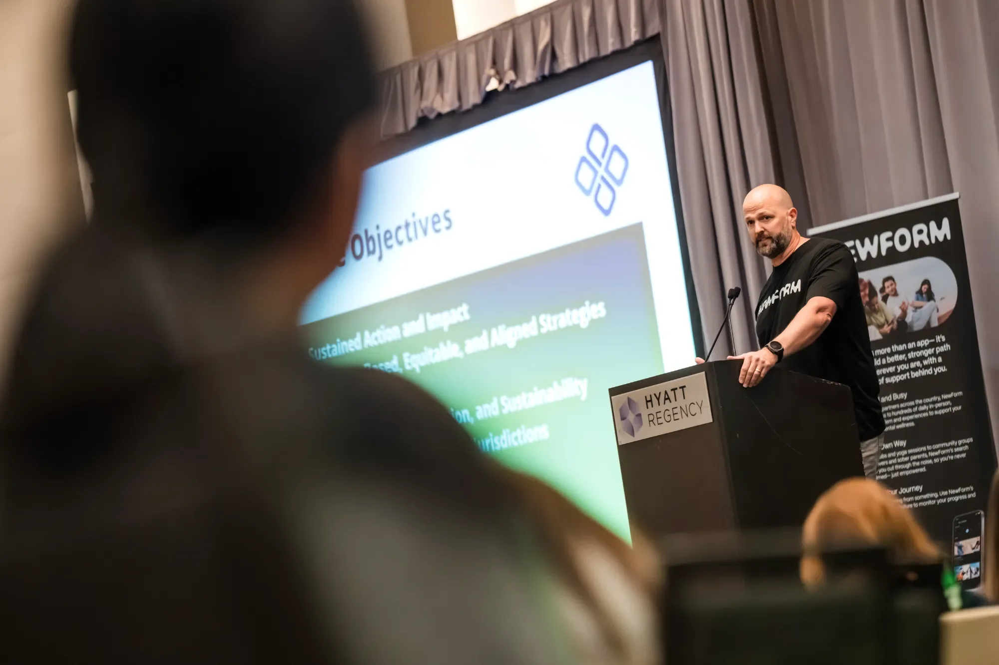 A man with a shaved head and beard stands at a podium with a "Hyatt Regency" sign, giving a presentation with a large screen behind him displaying the word "Objectives" and bullet points. There is a banner to his right with the word "NewForm" and an 