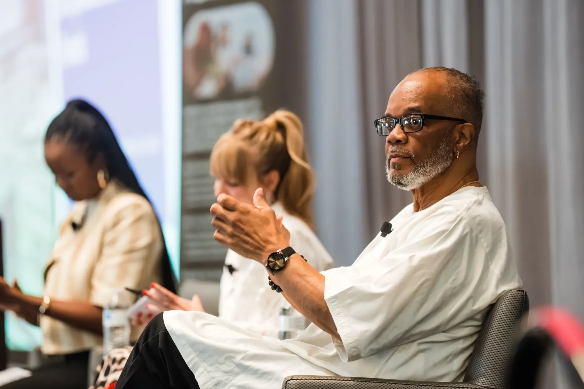 An elderly man with glasses and a beard sits attentively during a conference, with other people around him also listening.