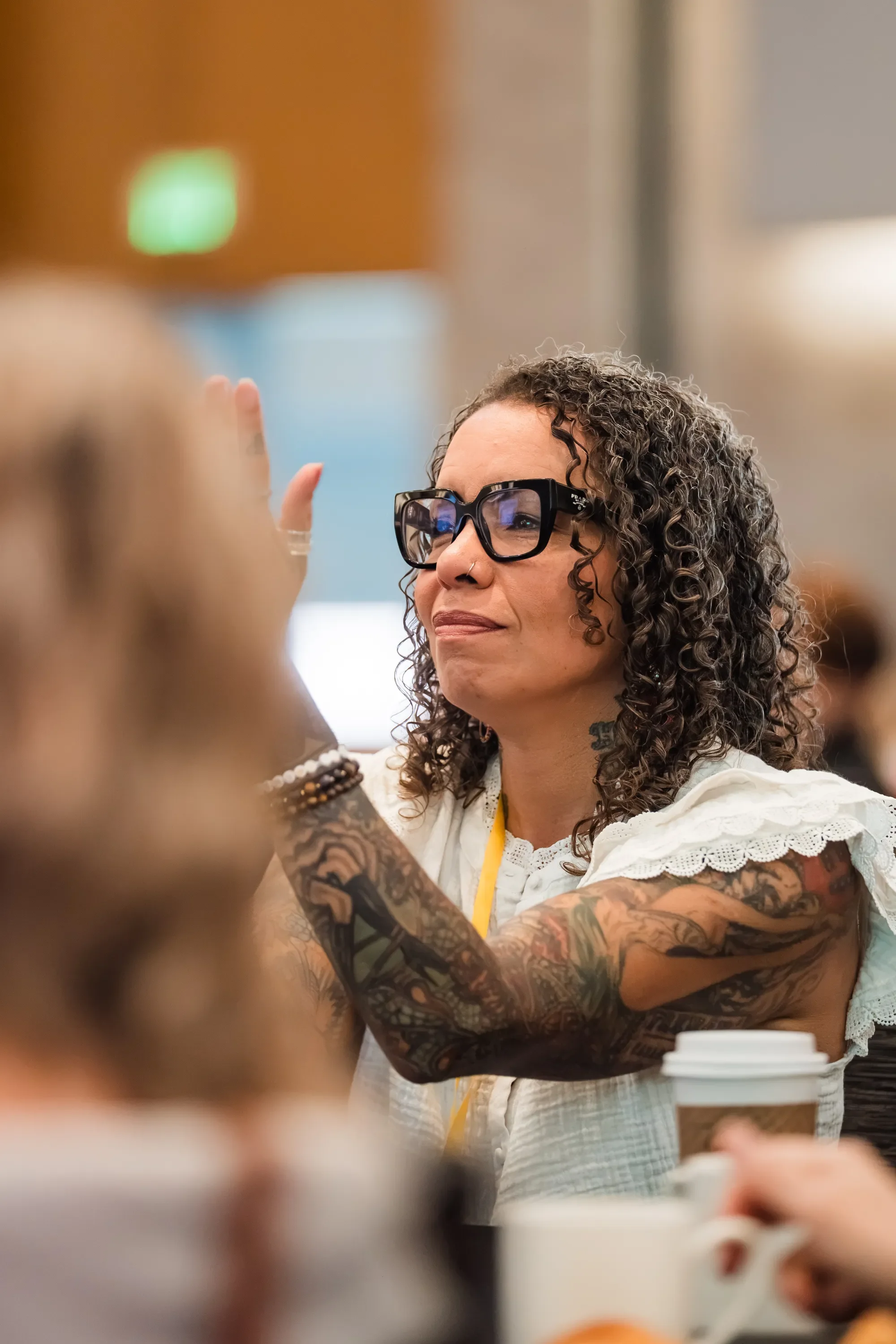 Woman with curly hair, glasses, and tattoos on her arms, smiling and gesturing with her hand in a crowded indoor setting.