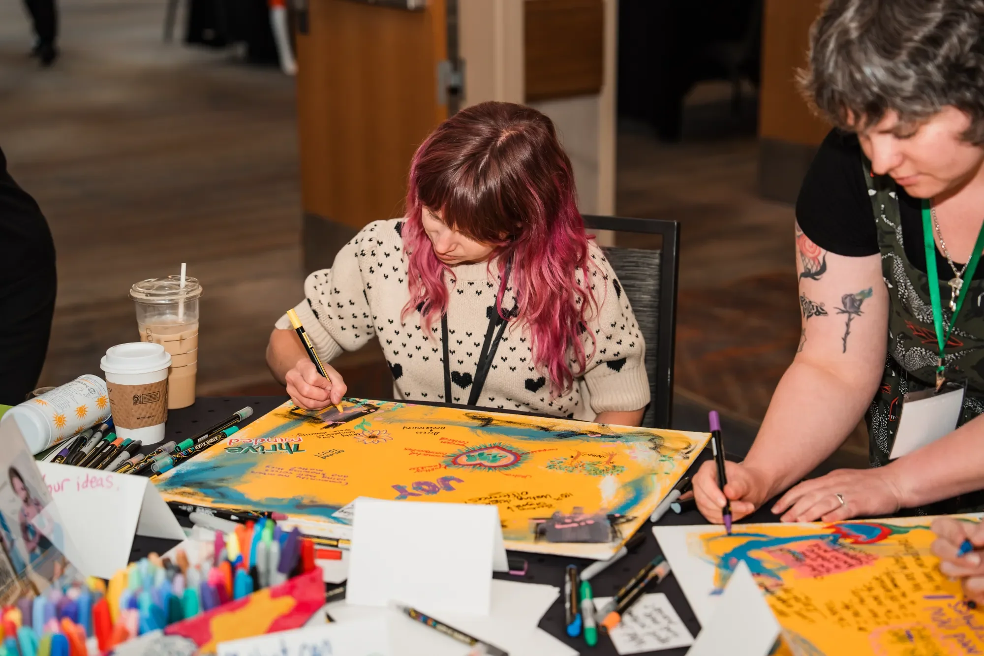 Two women sit at a table filled with colorful markers, pens, and a large yellow poster board, engaging in arts and crafts in a room with wooden floors and walls.