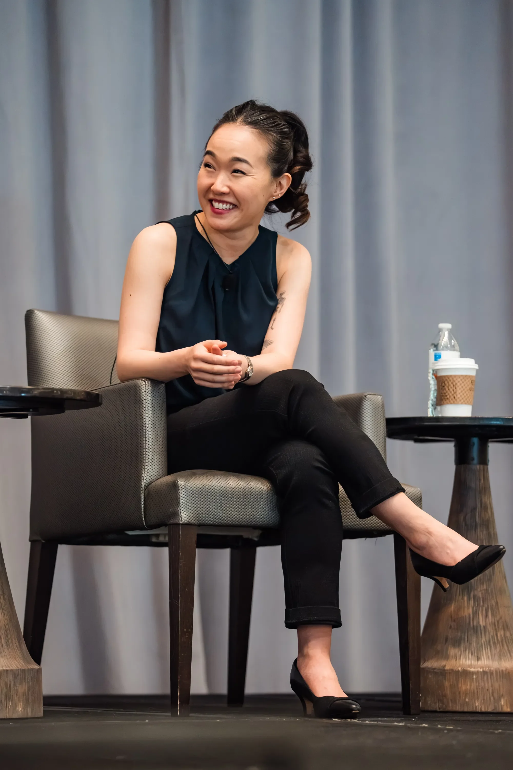 A woman with dark hair styled in a side ponytail, wearing a sleeveless black top and black pants, sitting on a chair with her hands clasped, smiling and looking to her right. There is a table with a water bottle and coffee cup on her right, and the b