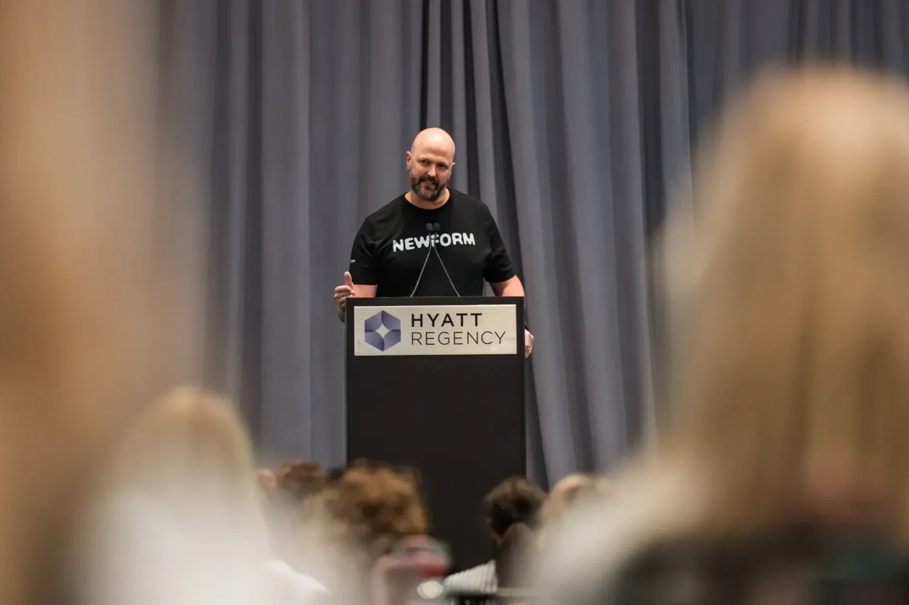 A man standing at a podium with a sign that reads 'Hyatt Regency' and a logo, wearing a black shirt with 'NEW FORM' written on it, speaking to an audience in front of a gray curtain.