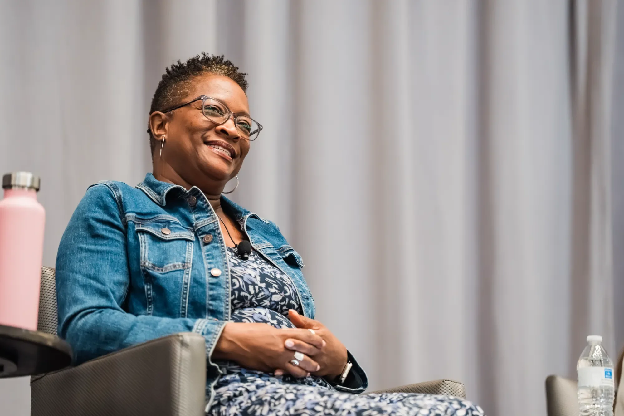 A woman with short curly hair, glasses, and hoop earrings smiling while sitting in a conference room. She is wearing a denim jacket over a patterned blouse, with a microphone clipped to her top. There is a pink water bottle to her left and a clear wa