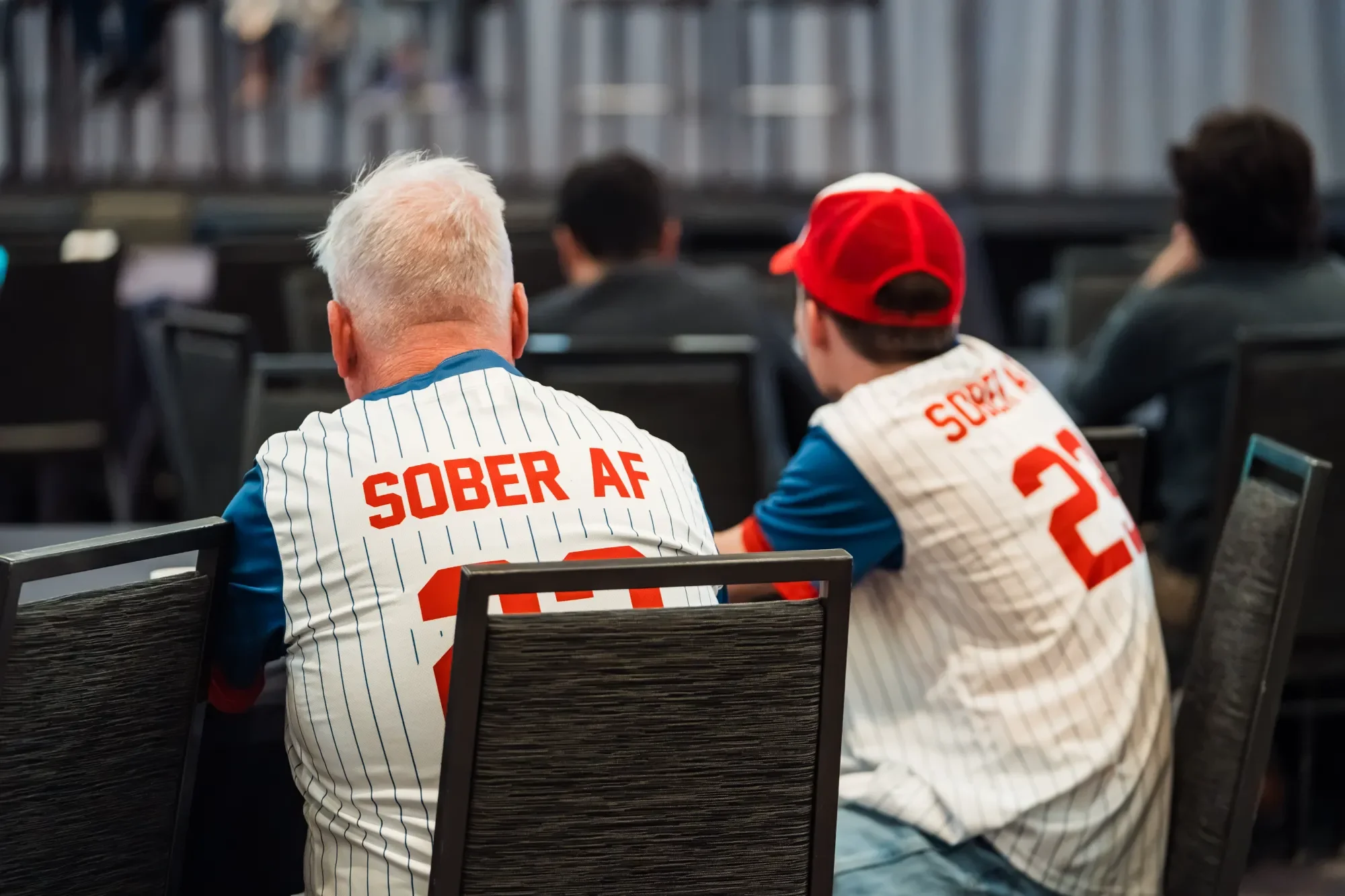 People sitting in an audience, wearing jerseys that say 'SOBER AF' on the back, in a conference room or auditorium setting.