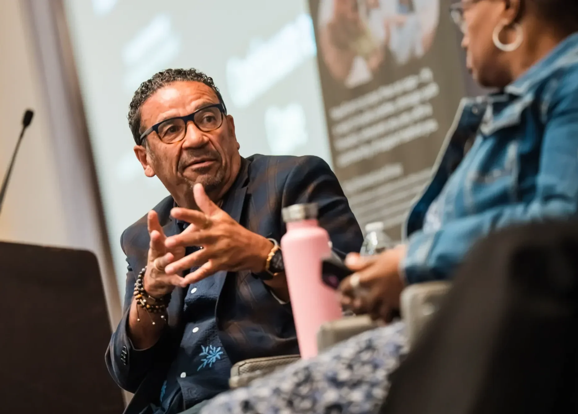 A man with glasses and bracelets is speaking to a woman with earrings, wearing a denim jacket, during a discussion at a table with various objects, including a pink bottle.