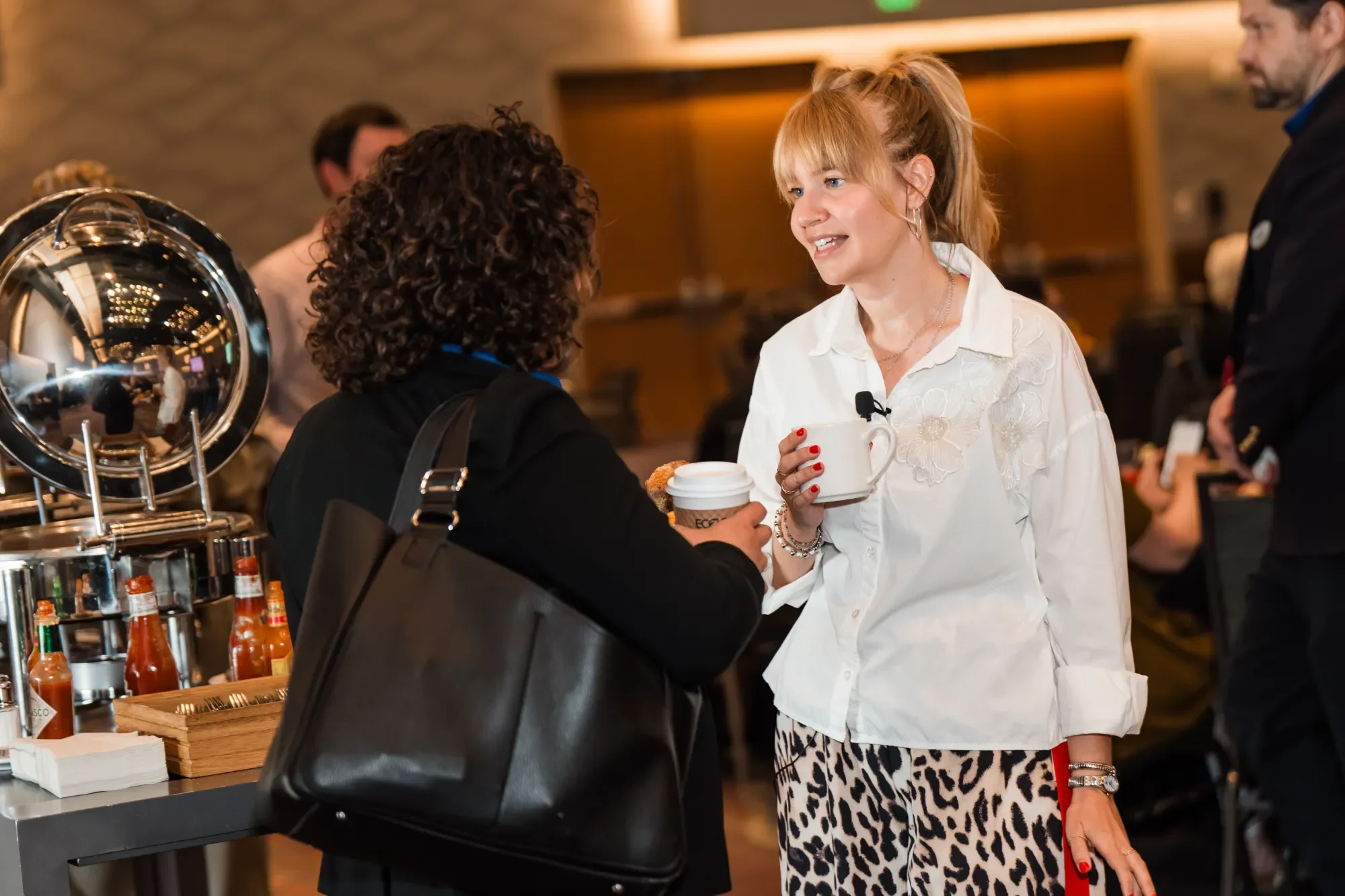 A woman with blonde hair and a white floral blouse is talking to another woman with curly dark hair and a black outfit at a social gathering. The woman with blonde hair is holding a white mug and a takeaway coffee cup while smiling. There are bottles