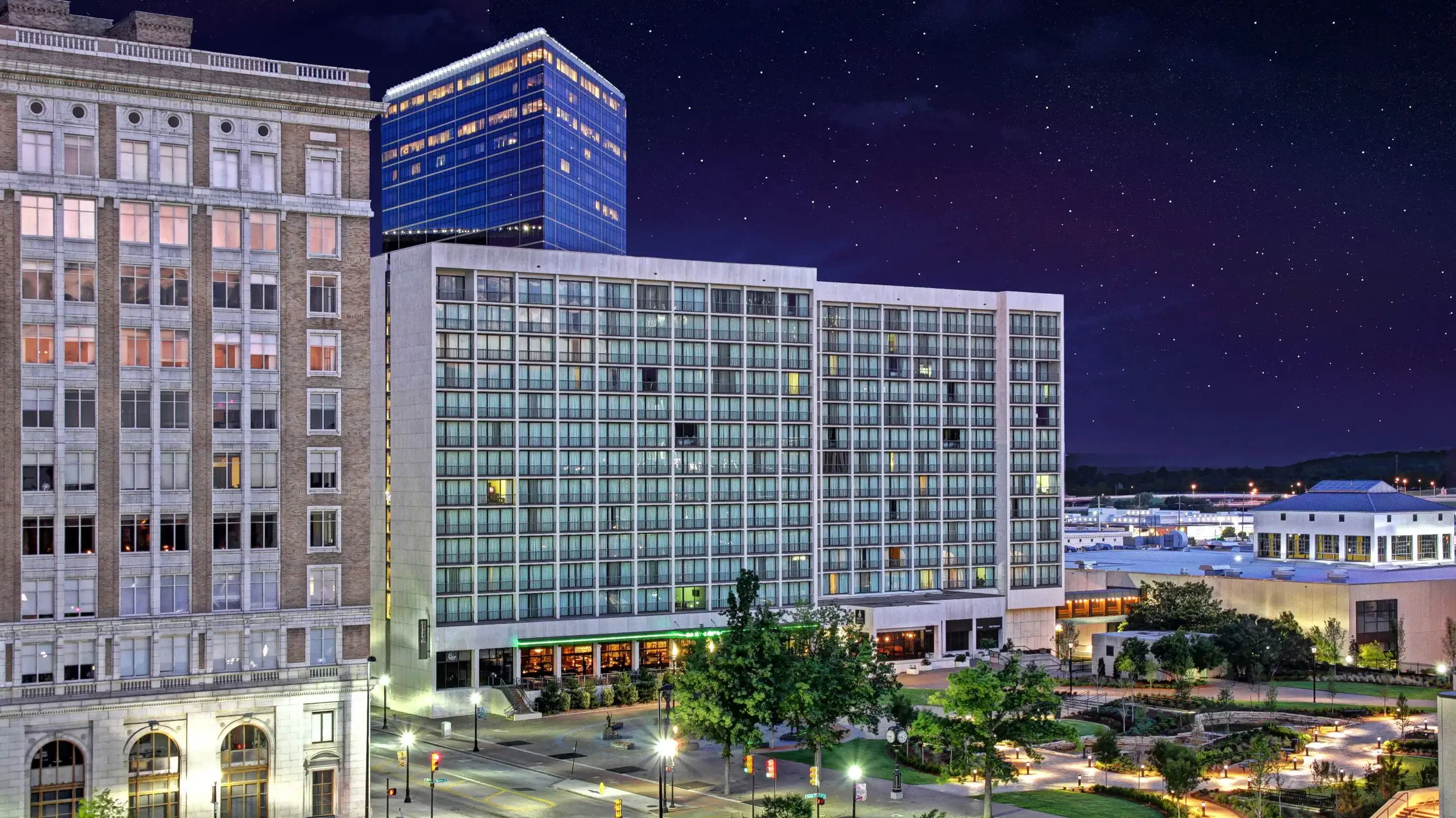 Nighttime cityscape showing several tall buildings with illuminated windows, a starry sky, and a park with trees and streetlights in the foreground.