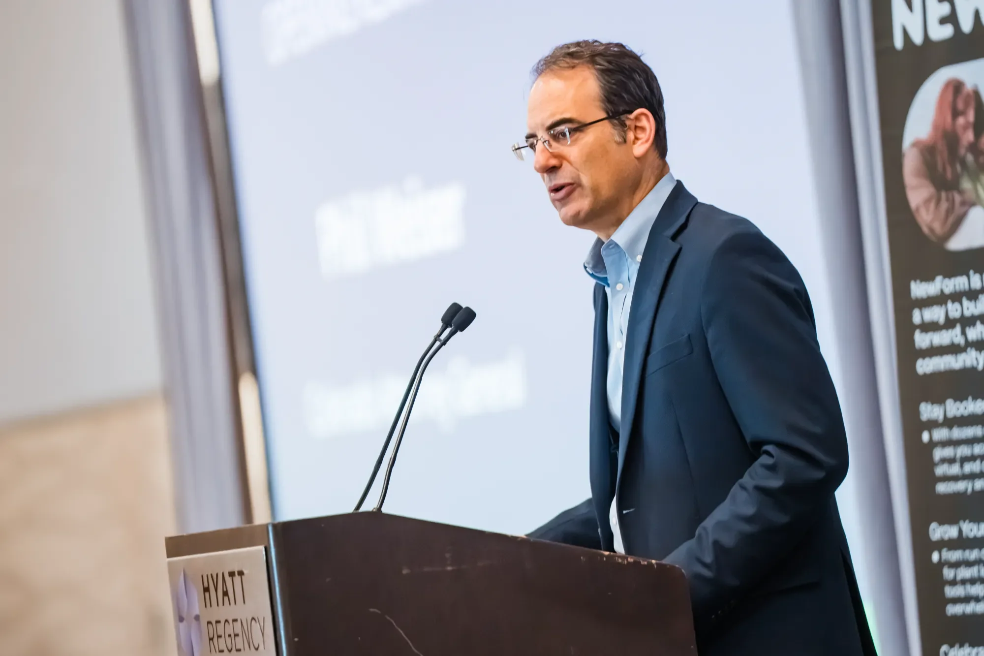 A man with glasses wearing a dark blazer and blue shirt giving a speech at a podium with the Hyatt Regency logo, in front of a large screen and a poster with text and a photo of a woman.