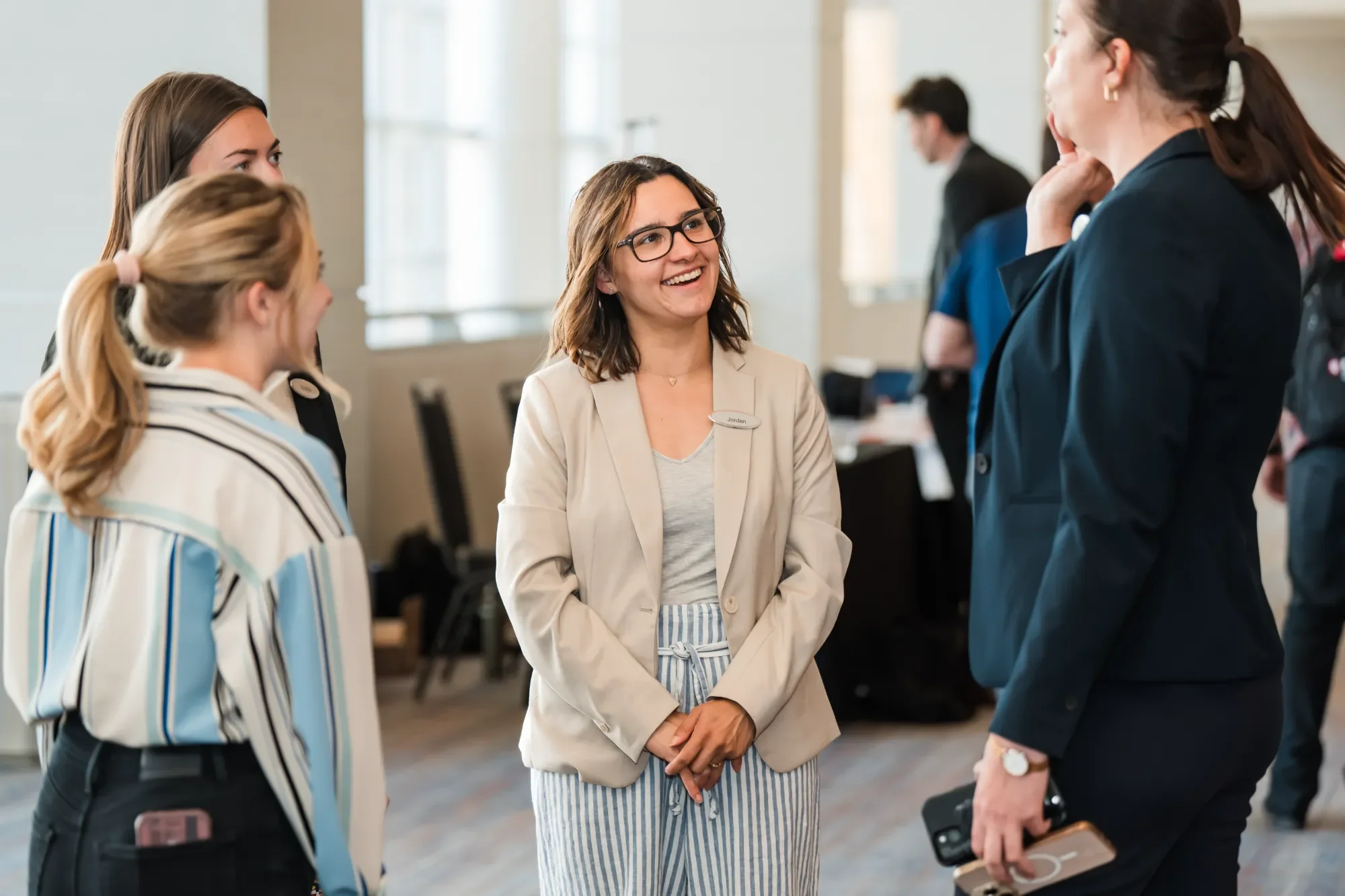 A woman in glasses and a beige blazer is smiling and talking to another woman with her hand on her chin. Two other women are in the foreground, one with a ponytail and striped blazer and the other holding a phone and glasses. In the background, a man