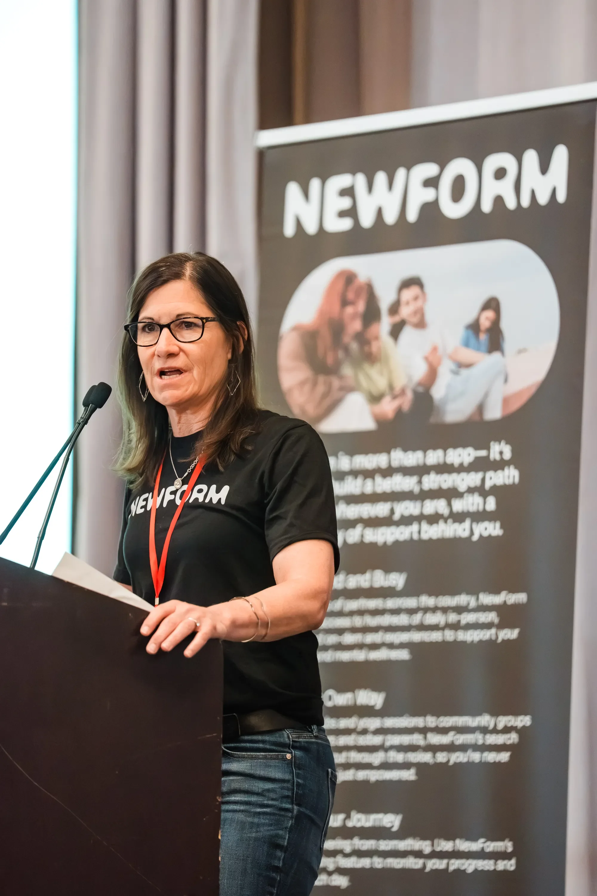 A woman wearing glasses and a black t-shirt with 'NEWFORM' written on it is speaking at a podium during a presentation. Behind her, there is a large banner with the same 'NEWFORM' logo and text related to community support and growth.