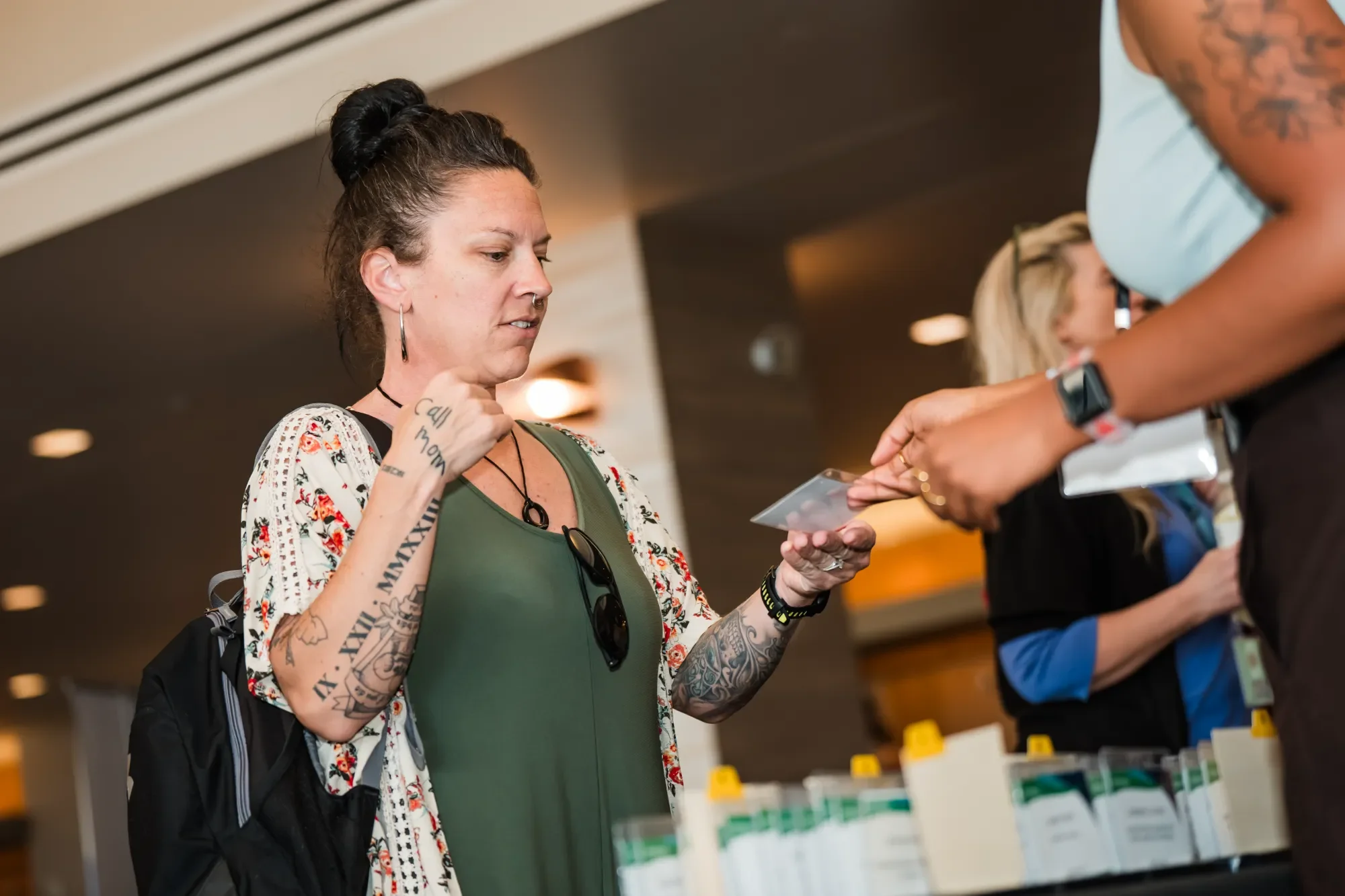 A woman with tattoos on her arms and a nose ring, wearing a green top and a floral kimono, is looking at her phone at a pharmacy counter. Multiple people are in line, and pharmacy shelves are visible in the background.