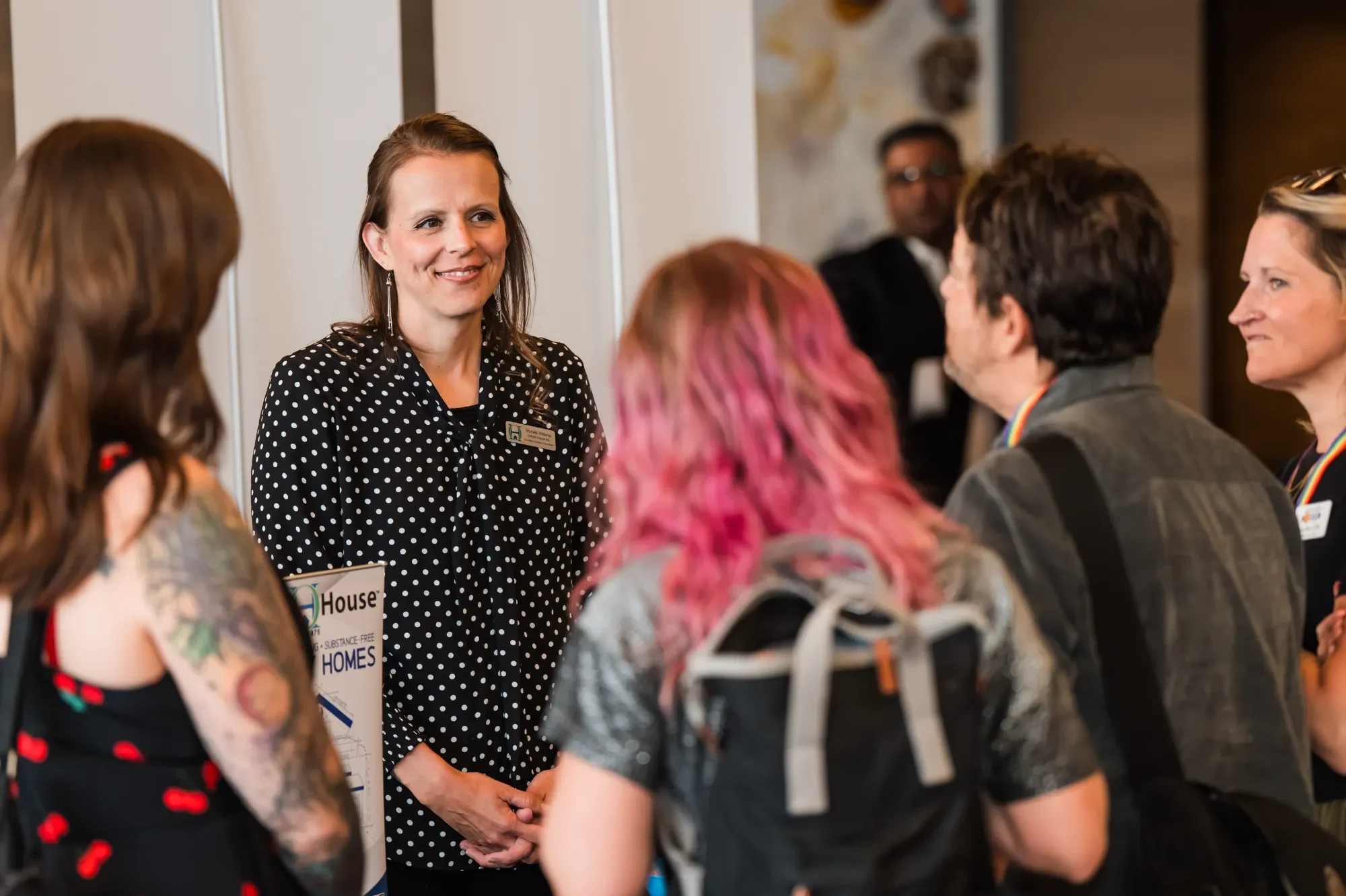 A woman with shoulder-length brown hair wearing a black and white polka dot shirt and earrings is smiling and talking to a group of people at an indoor event. The group includes individuals with colorful hair and tattoos, and some are wearing rainbow
