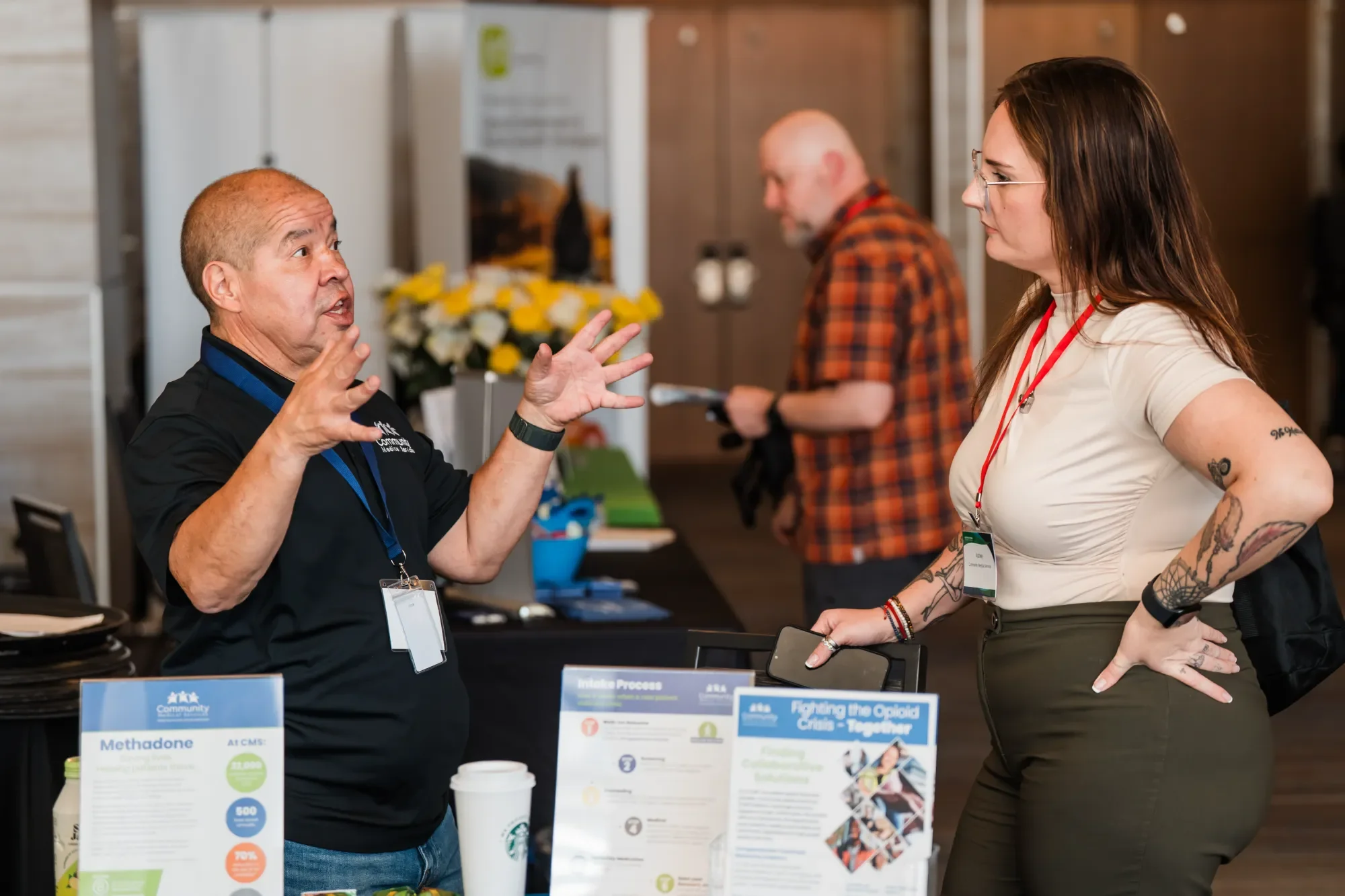 Two women and one man engaged in a conversation at an informational booth about opioid crisis prevention. The man is speaking and gesturing with his hands, while the women listen attentively. The booth displays posters and brochures related to community health and opioid treatment.