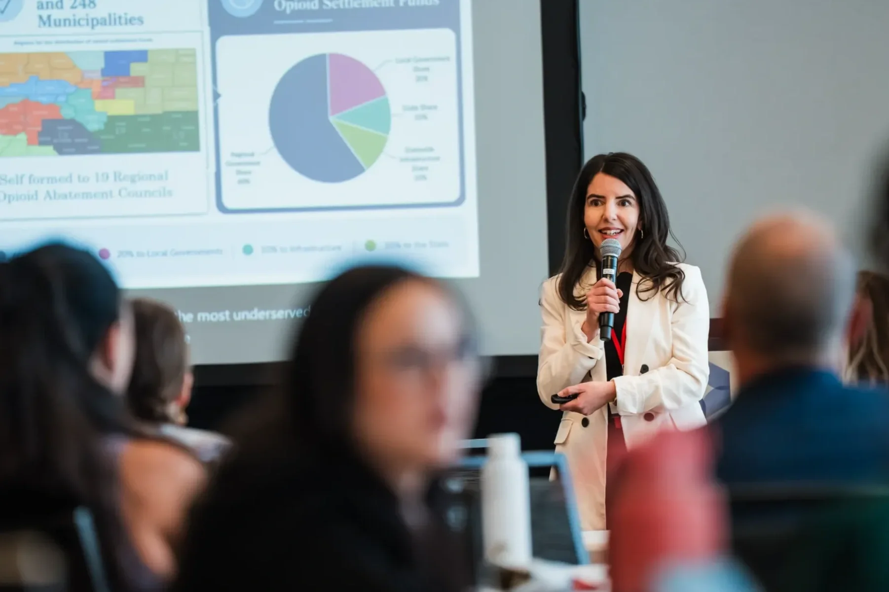 A woman with dark hair holding a microphone and a remote presents at a conference, standing in front of a projection screen displaying charts and a map, with an audience in the foreground.