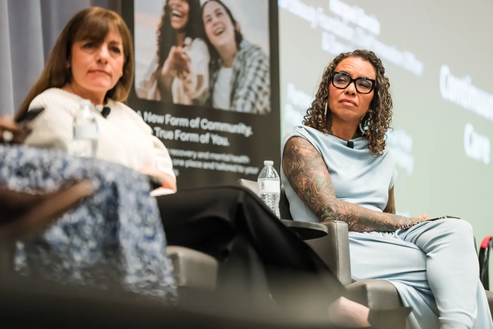 Two women sitting at a panel discussion or seminar, with one woman wearing glasses and a sleeveless top with tattoos on her arm, and a poster in the background showing a group of smiling women and text.