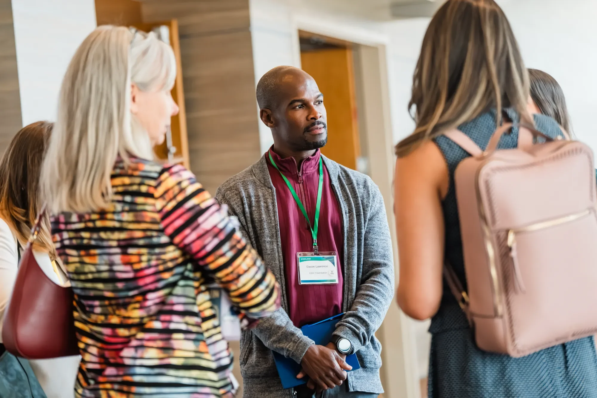 A group of diverse people attending a conference or seminar, with one man in the center wearing a gray jacket and a name badge, listening to a speaker or presentation.