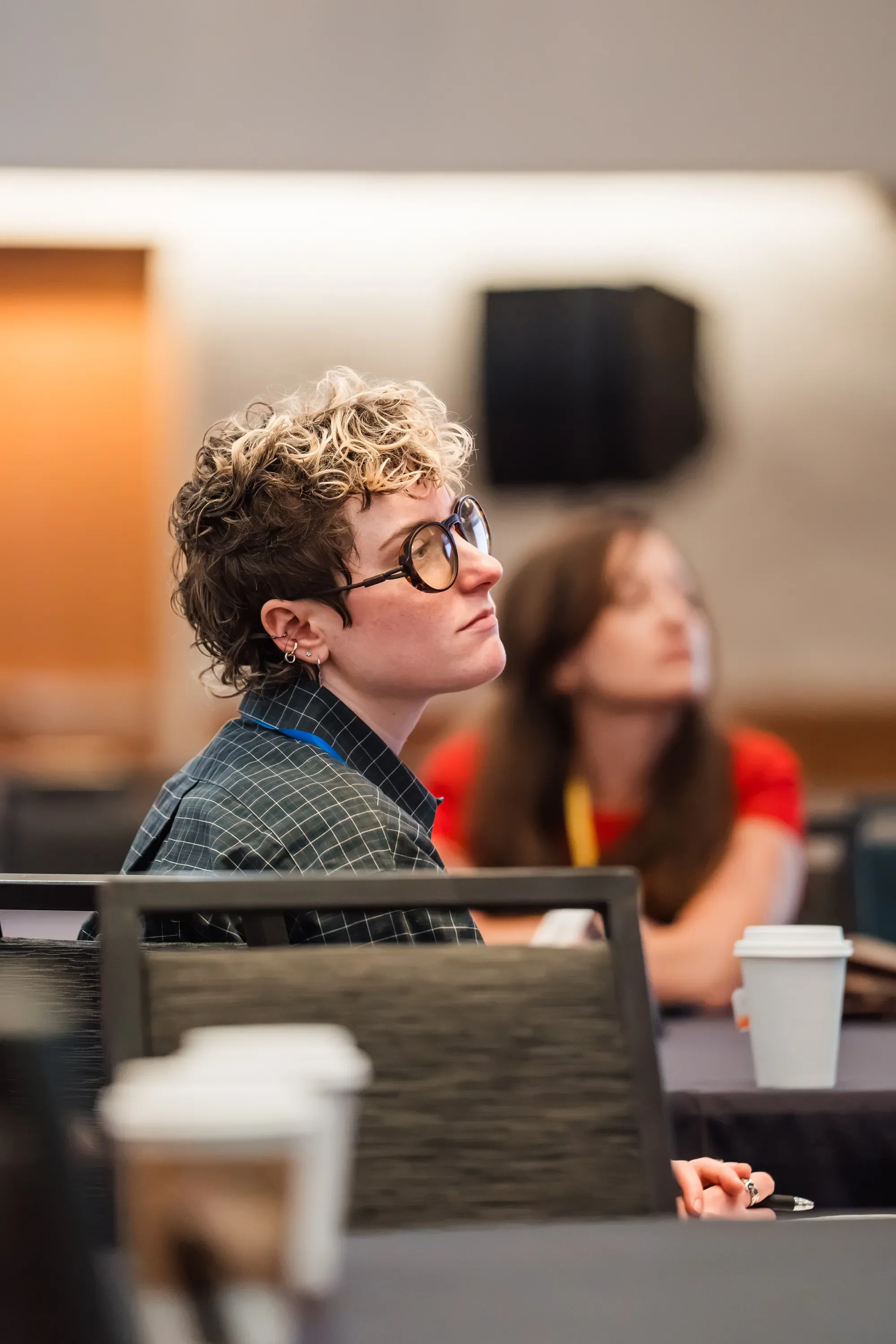 A woman with curly blonde hair, wearing glasses and a checked blazer, is sitting at a desk in a meeting or conference room. There is a woman with long brown hair in the background. The scene includes a table with a disposable coffee cup and some offi