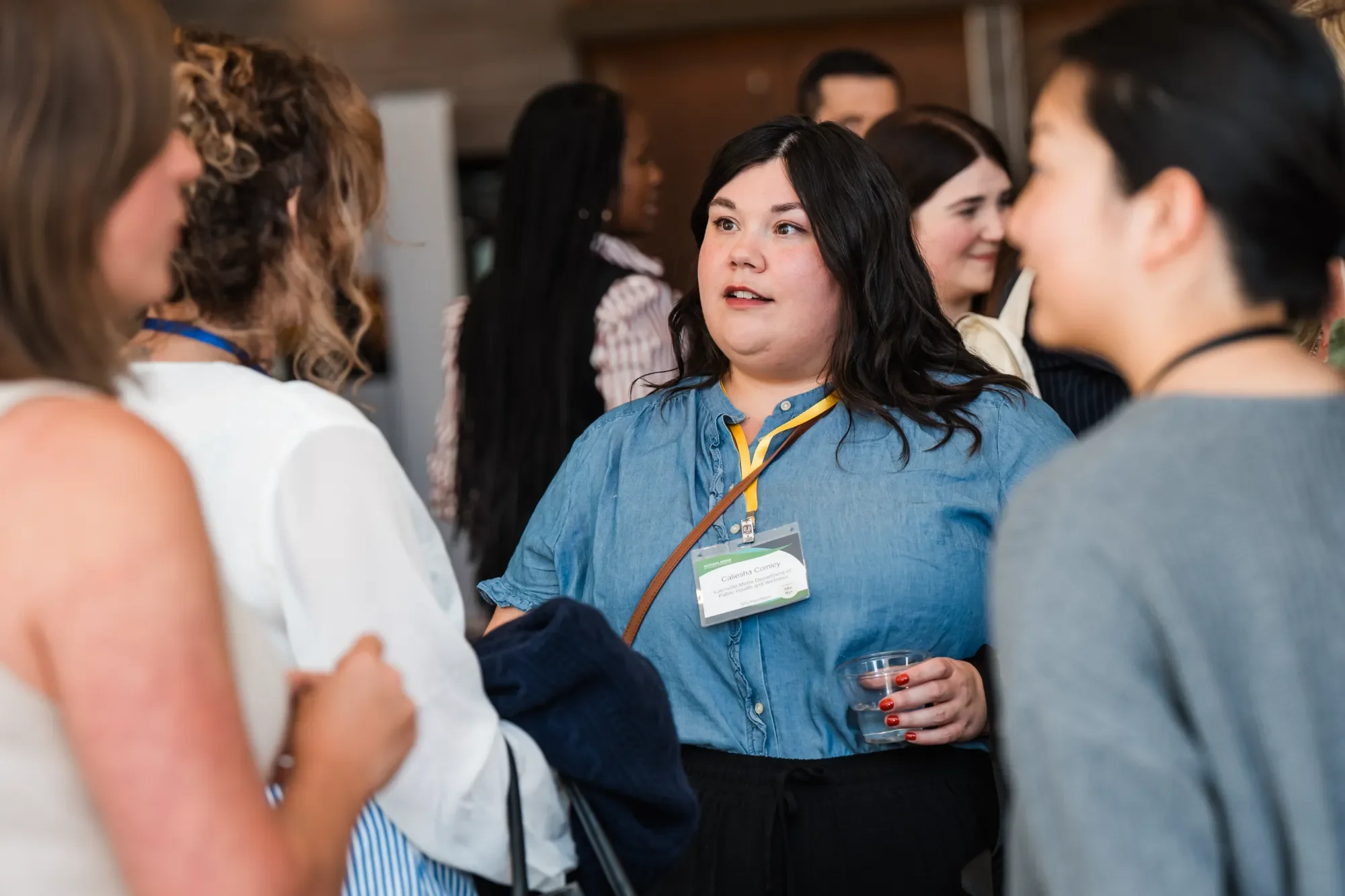 A group of women engaged in conversation at a social event, with one woman in focus wearing a blue shirt and a name tag, holding a drink, and talking to others.