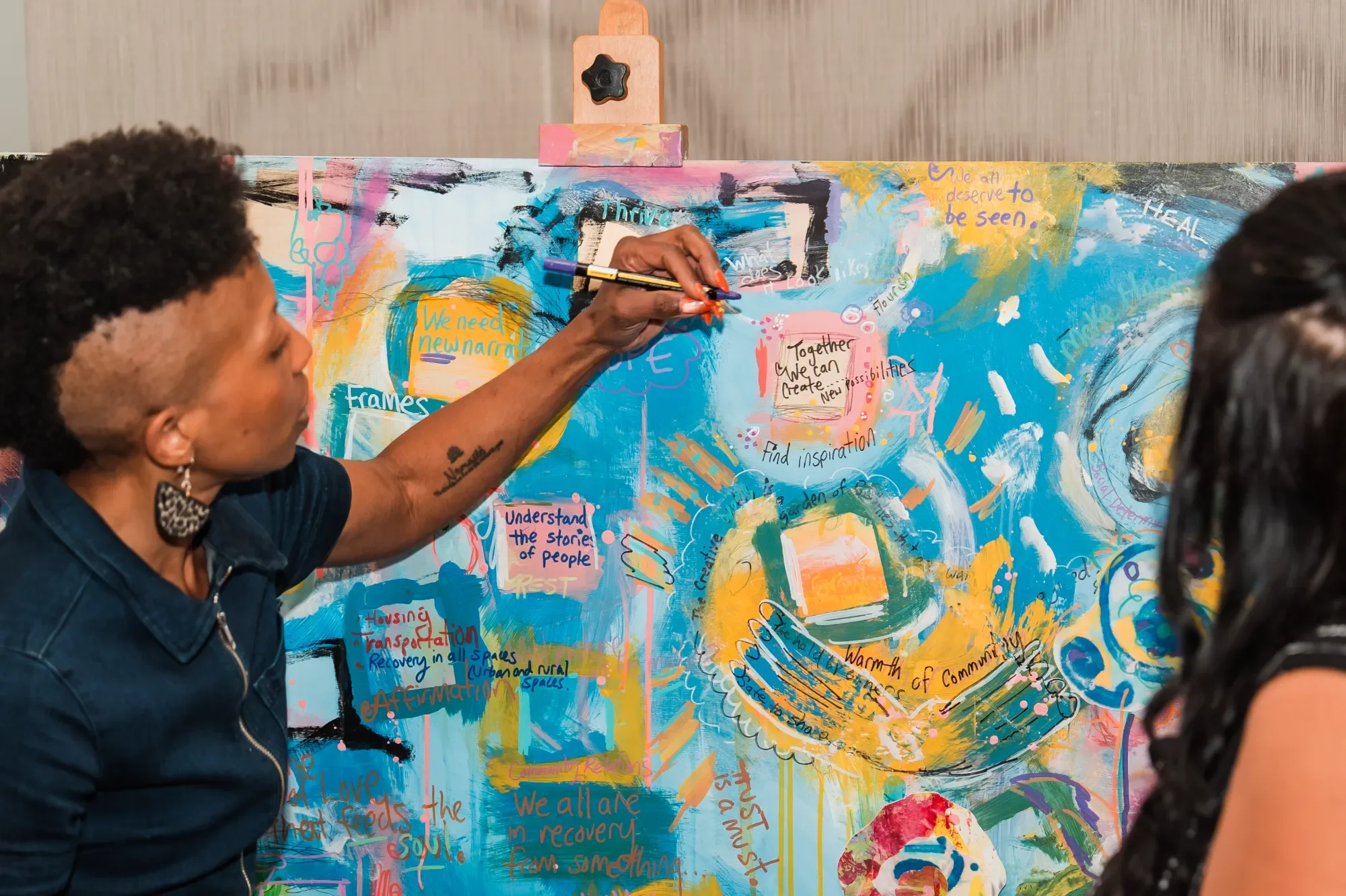 Close-up of a woman with short curly hair painting on a colorful, abstract mural with various handwritten messages and doodles, surrounded by a group of people.