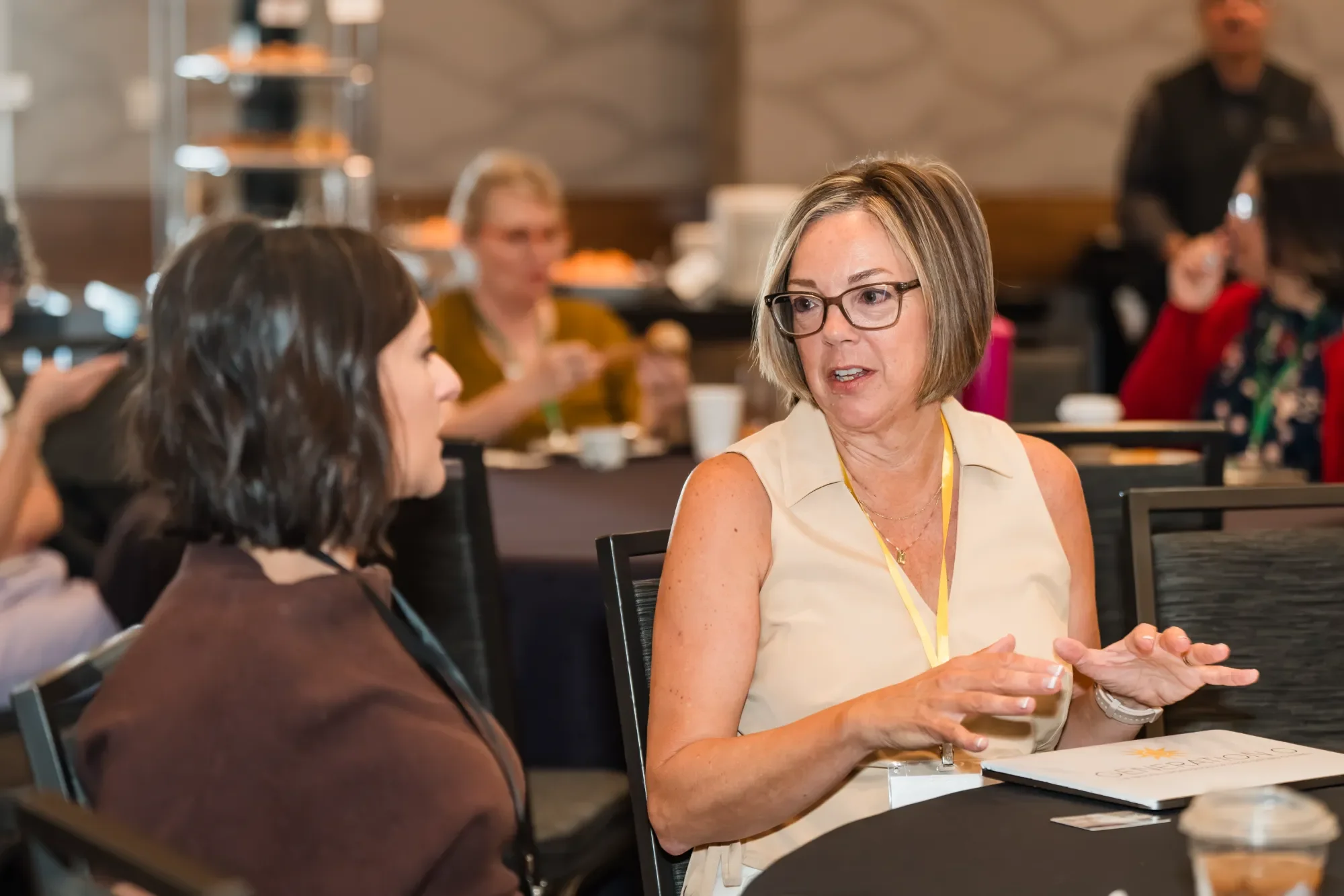Women seated at a conference table engaging in conversation, with other attendees in the background. One woman with glasses and short hair is talking, while another with dark hair listens.
