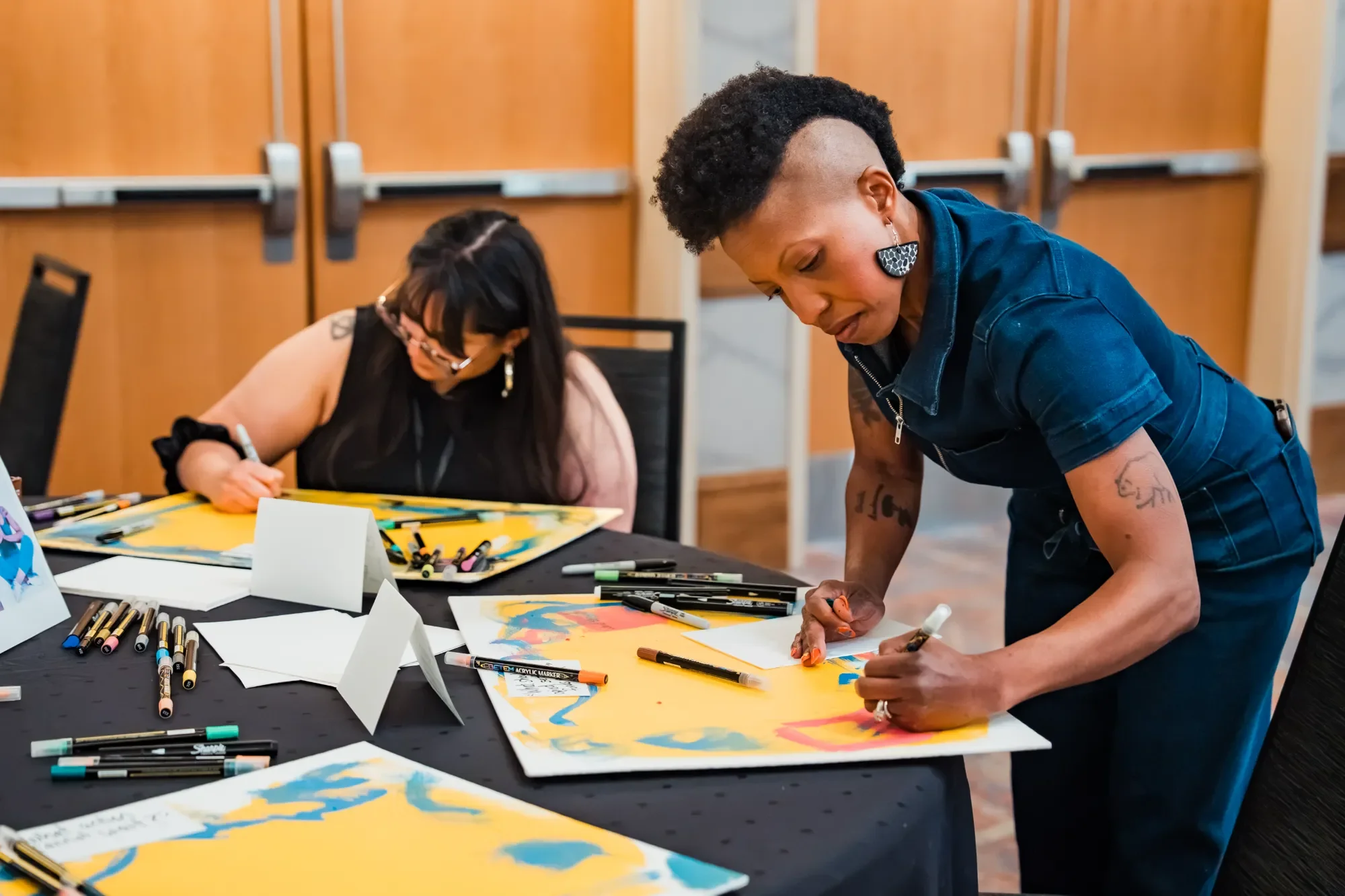 Two women are engaged in an art activity at a table filled with markers and drawings. One woman with short curly hair is leaning over, drawing or writing on paper, while the other woman with long straight hair is sitting and writing or drawing on her