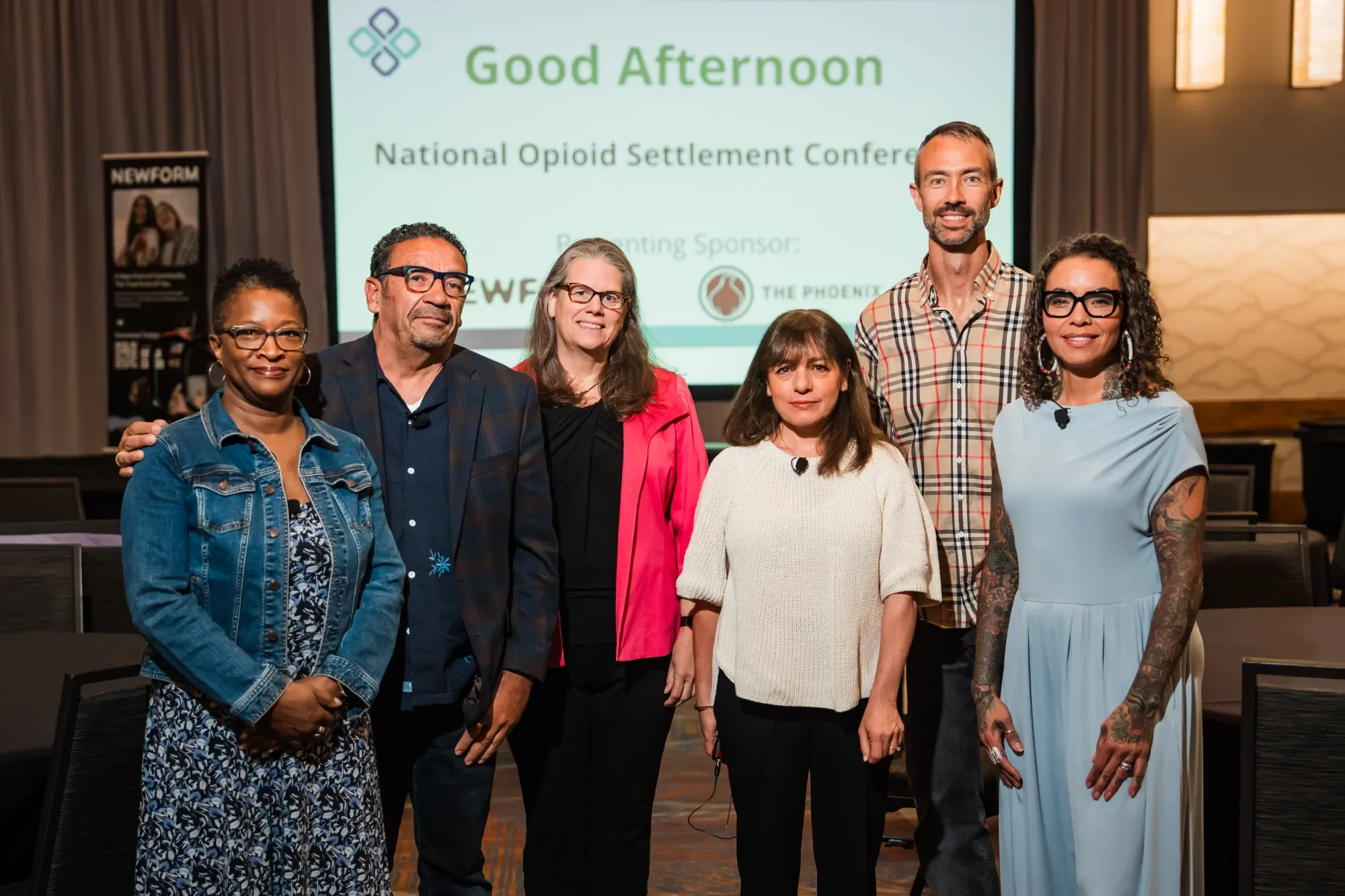 NOSC team standing together at a conference, with a presentation screen in the background displaying the words "Good Afternoon" and "National Opioid Settlement Conference."