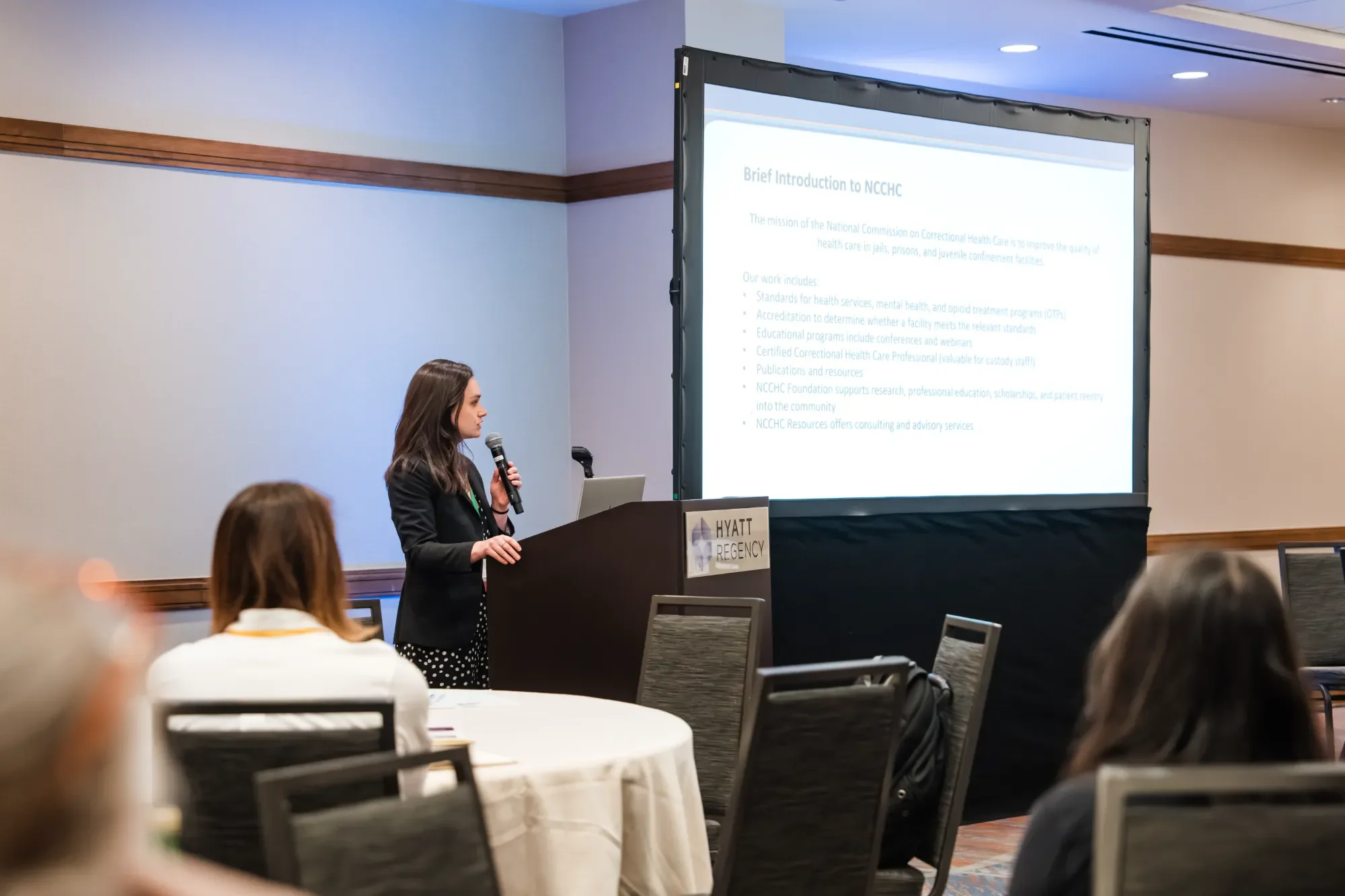 A woman is giving a presentation at a conference in a hotel conference room. She is standing at a black podium labeled 'HYATT REGENCY' while holding a microphone. Behind her, a large screen displays a slide titled 'Brief Introduction to NCCHC' with b