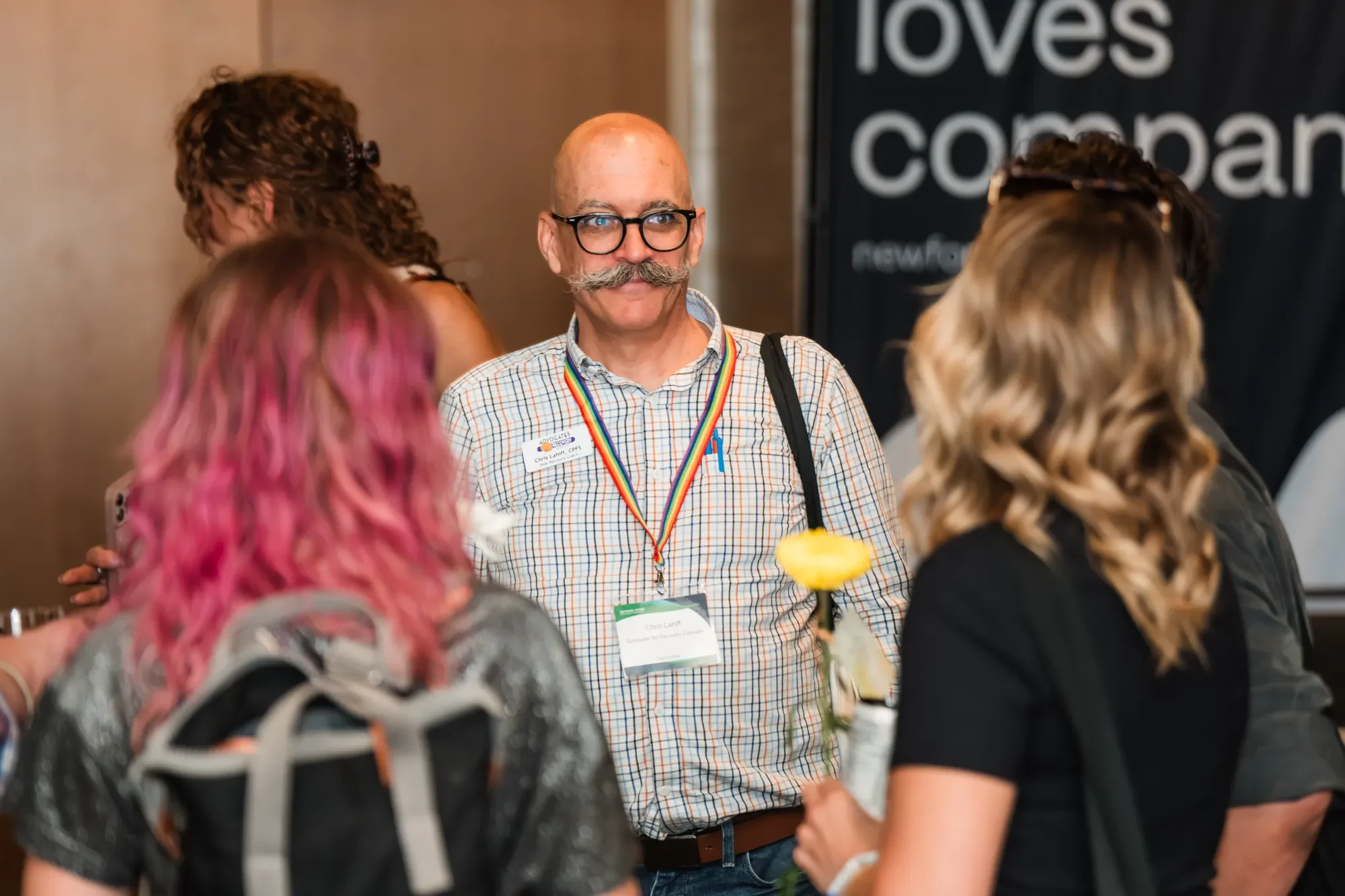 A man with a red beard and glasses, wearing a checkered shirt and a rainbow ribbon lanyard, standing among a group of women with various hair colors and styles at an indoor event.