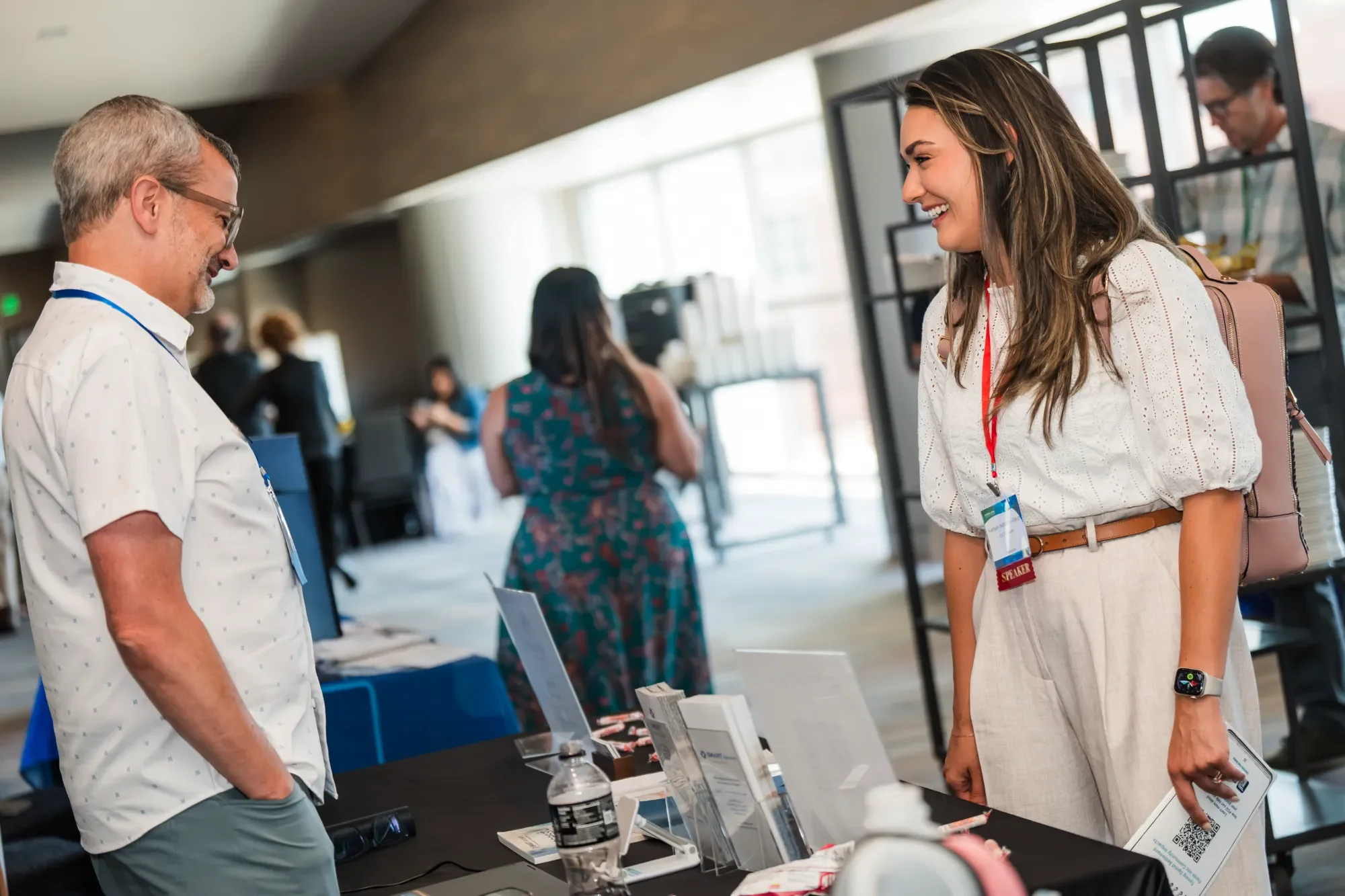 A man and woman are smiling at each other while standing at a conference or event booth. The woman is wearing a white dress, a conference badge, and a smartwatch. The man is wearing glasses, a white shirt with small pattern prints, and has a conferen