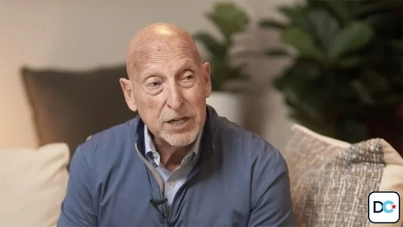 An elderly man with a bald head and a beard, wearing a blue jacket, is sitting on a couch in a living room with decorative pillows and plants in the background.