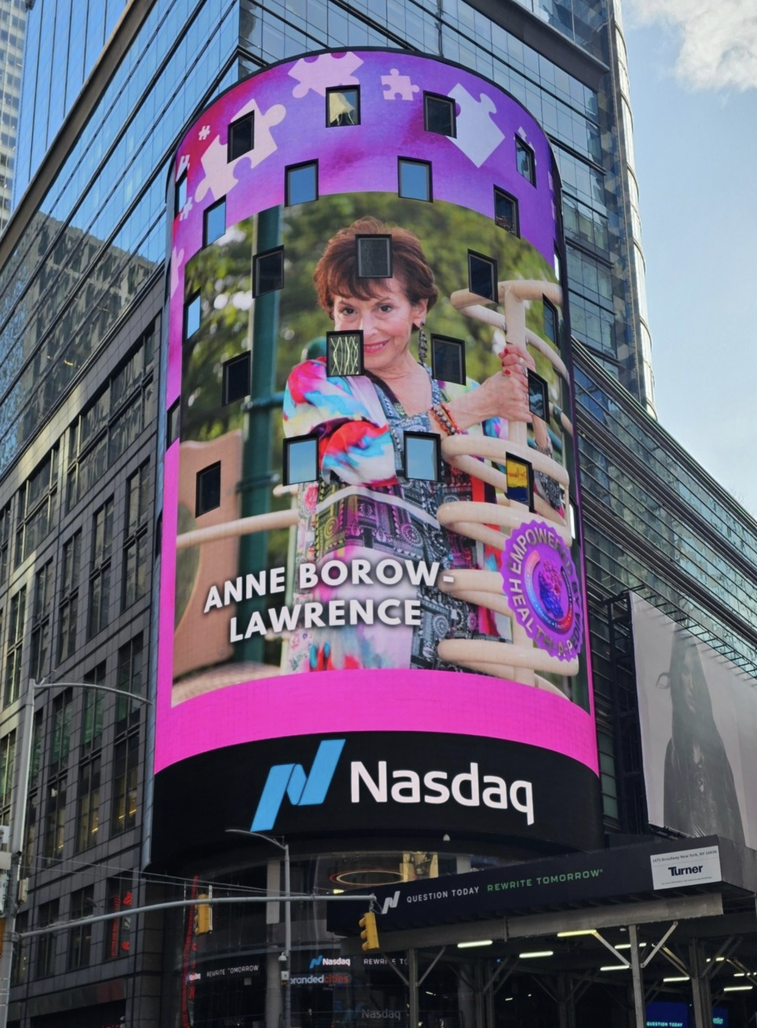 Digital billboard on a Nasdaq building featuring a woman, Anne Borow-Lawrence, with a colorful outfit, standing in front of a playground ladder, with the words "Anne Borow-Lawrence" and a logo that promotes empowerment and health. The Nasdaq logo is at the bottom of the screen.