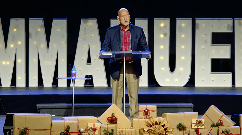 A man in a blue blazer and red plaid shirt stands at a clear podium on a stage with large illuminated letters spelling 'IMAGUE' behind him. The stage is decorated with wrapped gifts and holiday-themed ornaments, including Christmas baubles and ribbons.