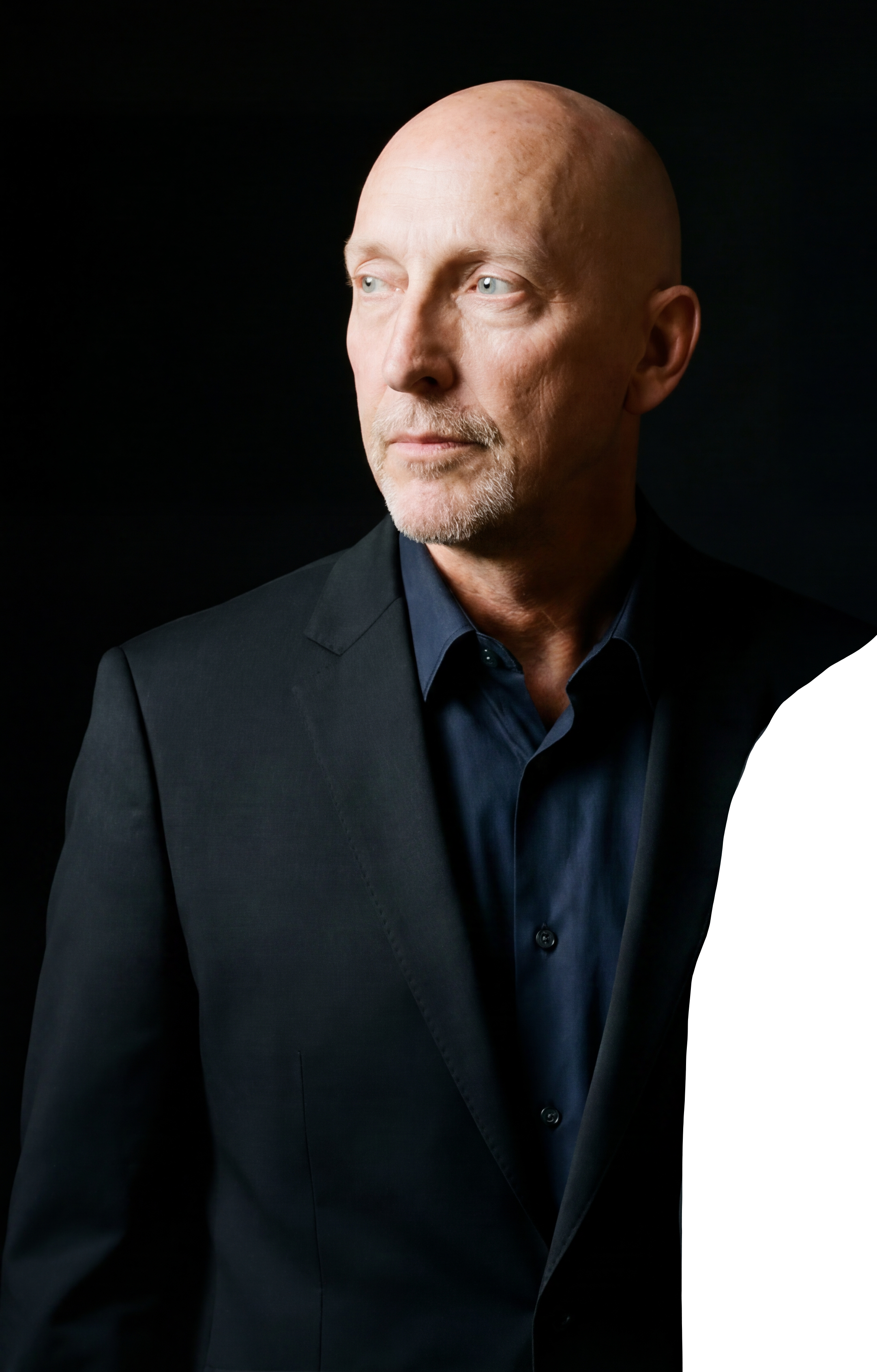 A middle-aged man with a shaved head and light complexion looking to the right, wearing a dark suit and shirt, against a black background.
