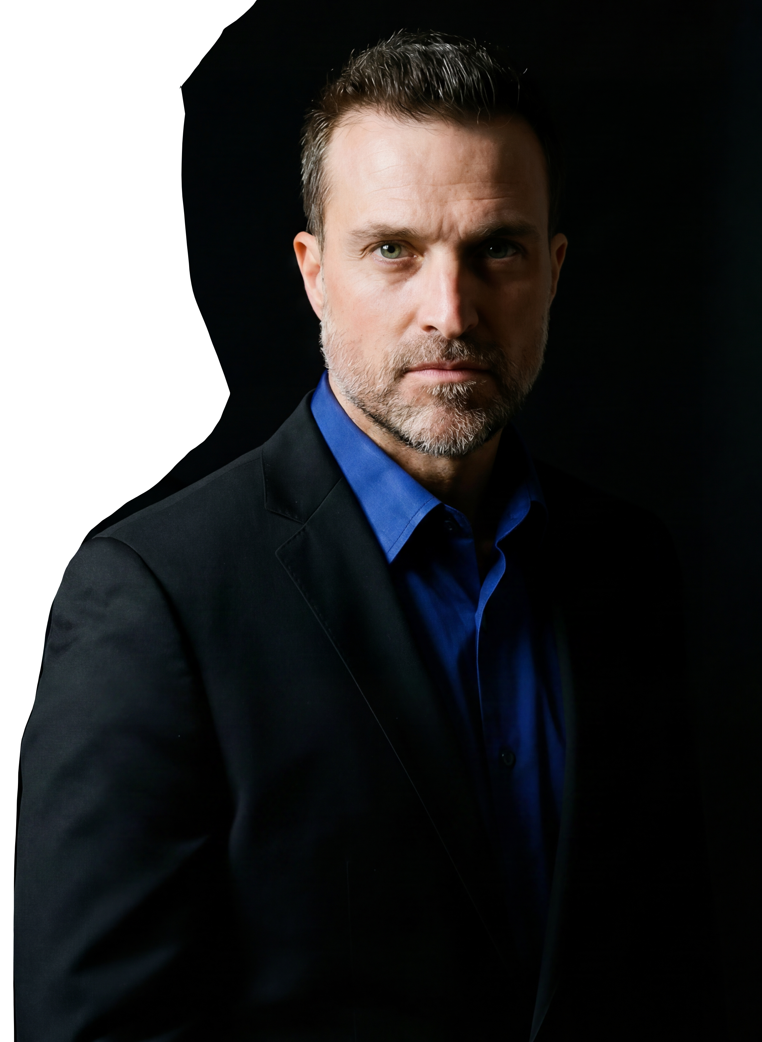 A man with short, dark hair, a beard, and green eyes wearing a black blazer and a blue dress shirt, looking directly at the camera against a black background.