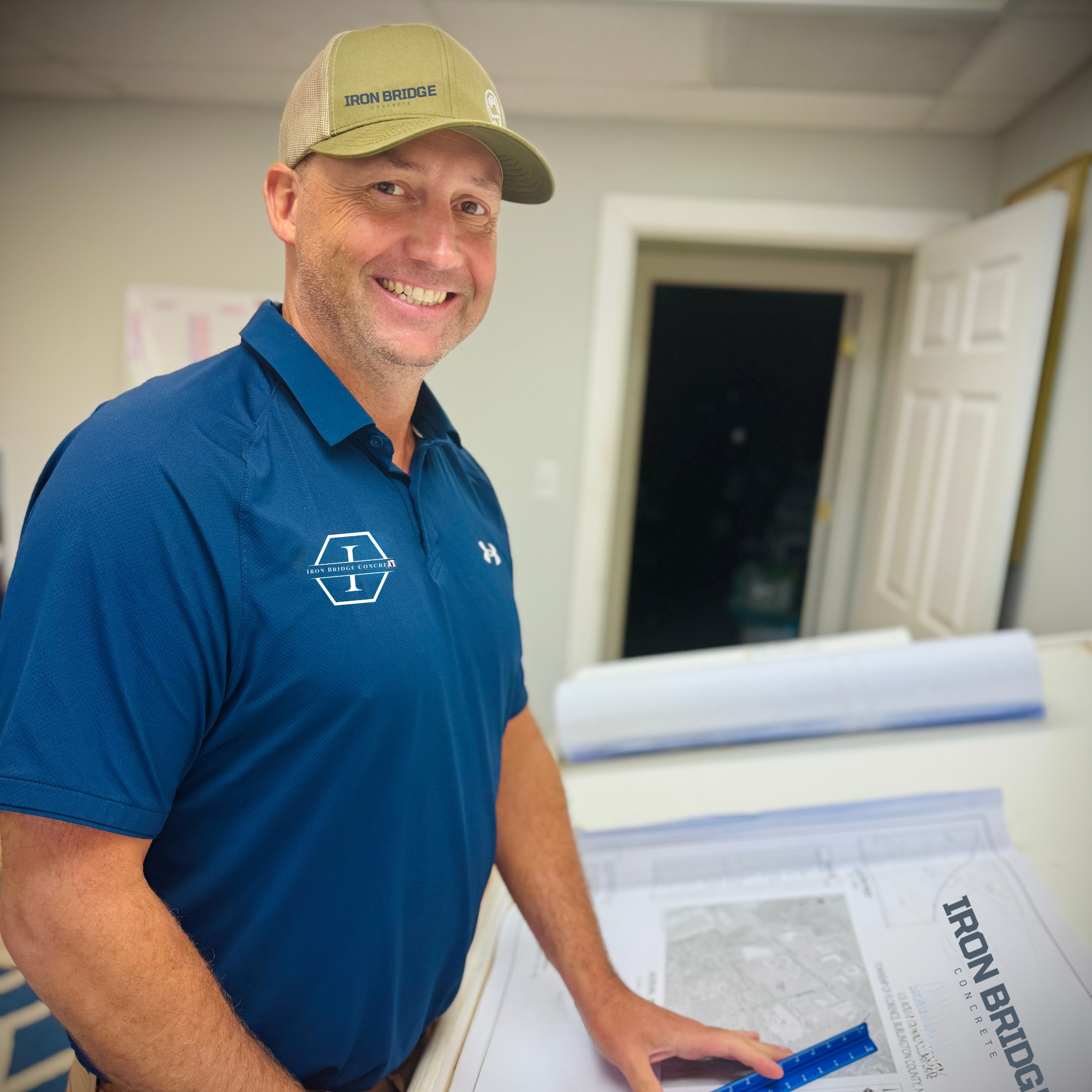 A man smiling wearing a beige baseball cap with "Iron Bridge Concrete" logo and a blue polo shirt with the same logo, working on a large construction blueprints or plans in an indoor setting.