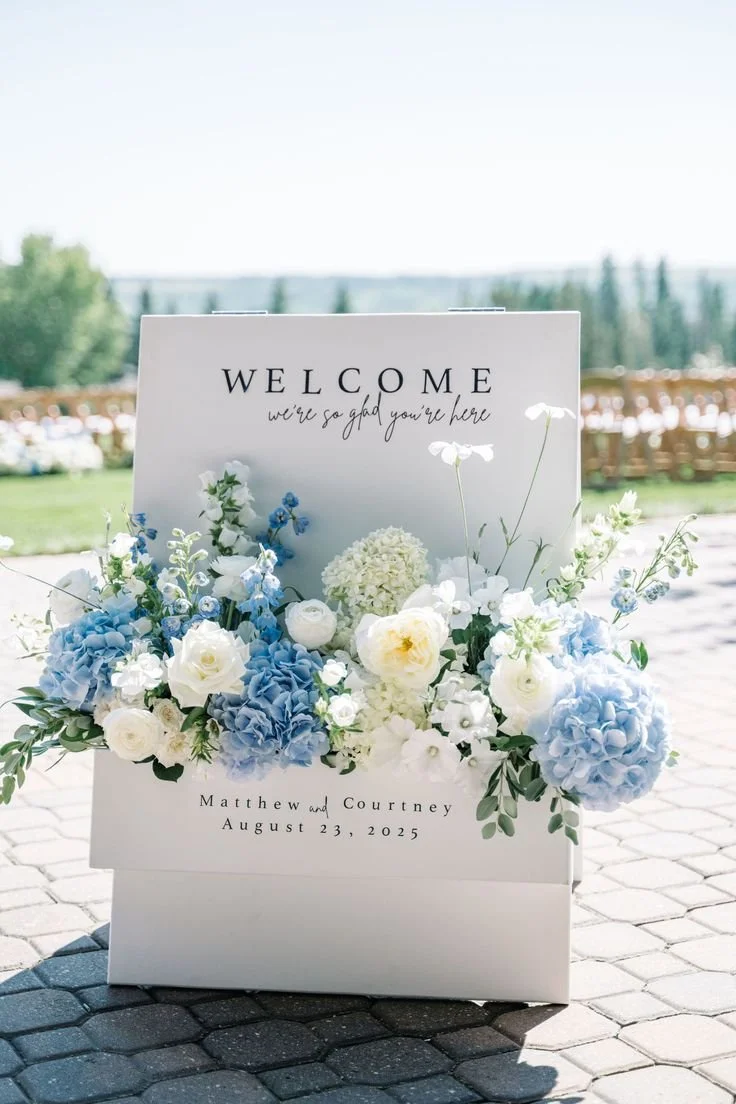 An A-frame White Welcome sign with pocket floral arrangement for a wedding ceremony, displaying the names of the bride and groom.