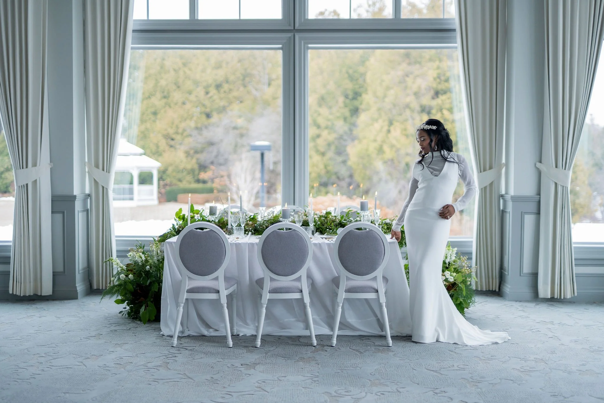 Elegant dining room set for a wedding with a white tablecloth, floral centerpiece, and candles, set before a large window with white curtains, overlooking trees outside. A woman in a white dress with a decorative headpiece stands beside the table.