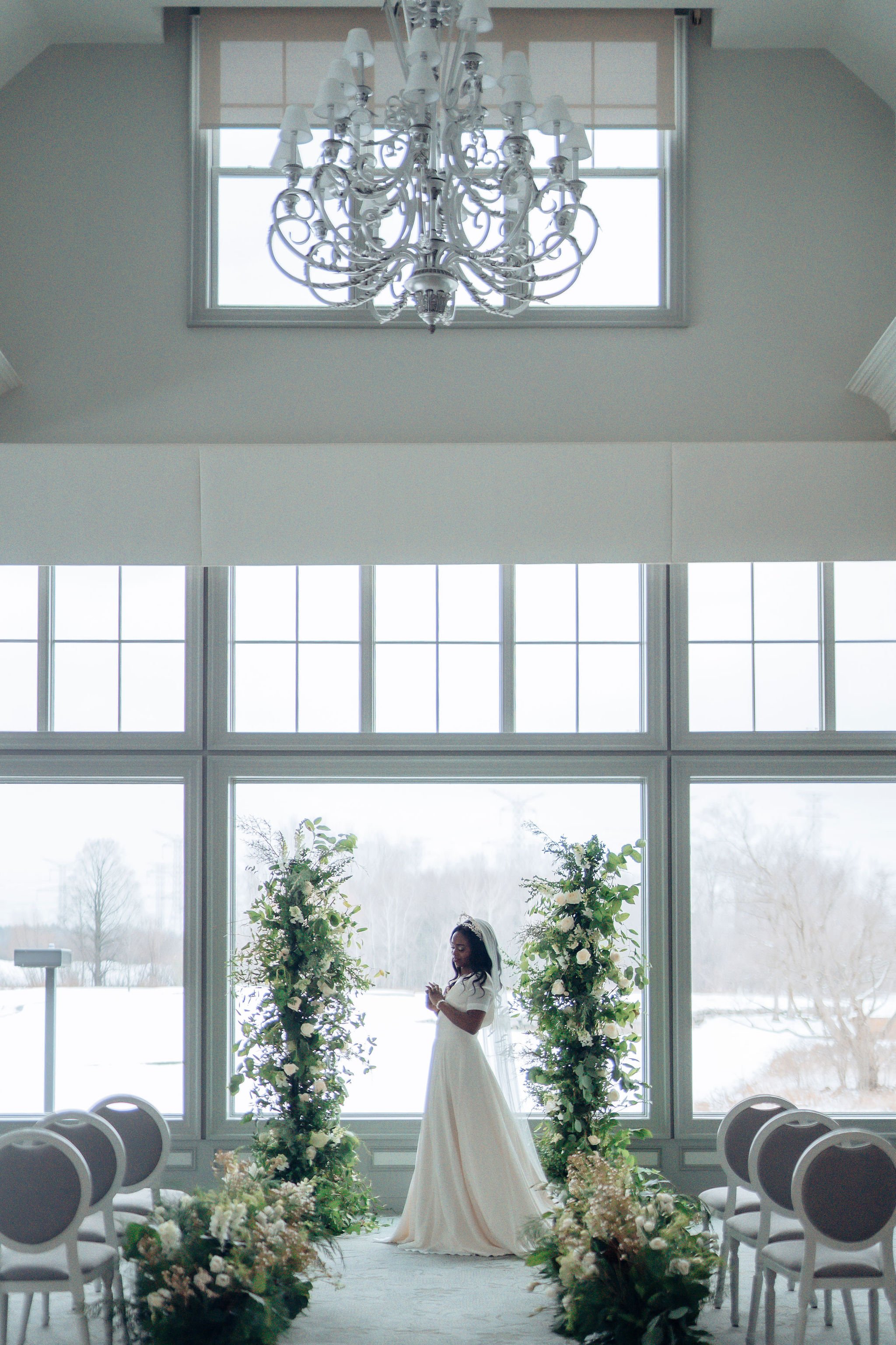 A woman in a wedding dress standing in front of a large window, with floral arrangements on either side, during a wedding ceremony in a bright, spacious room with a chandelier hanging from the ceiling.