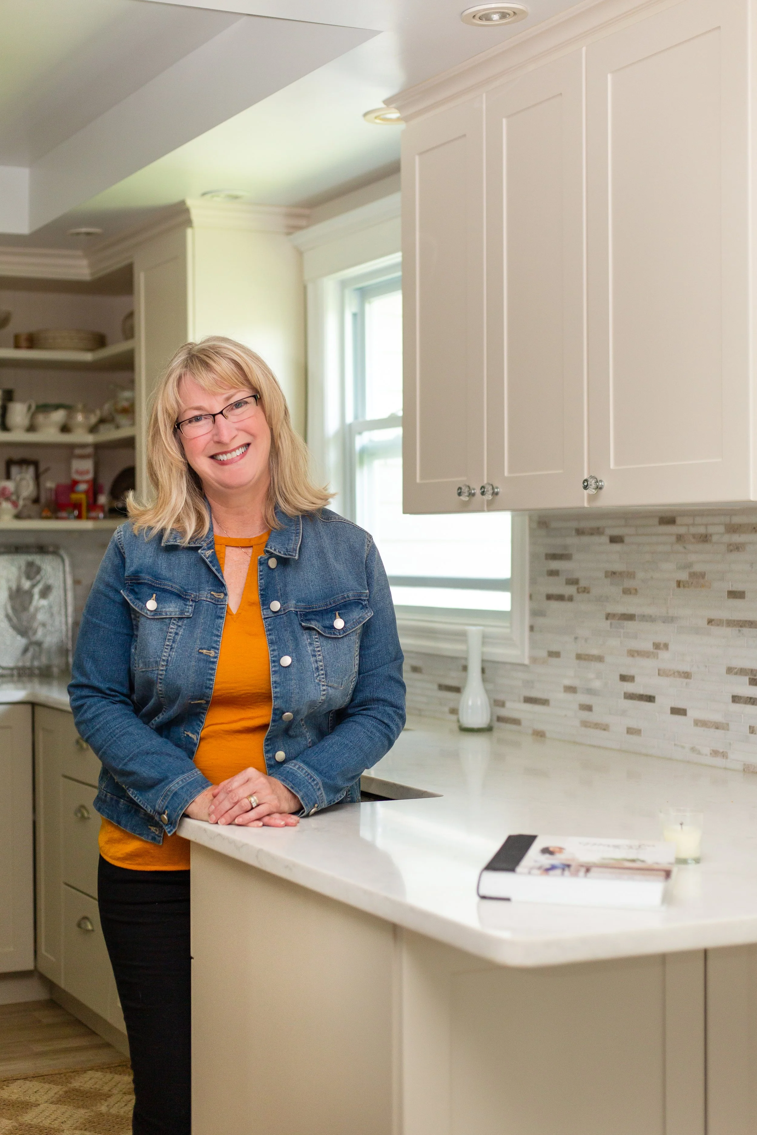 A woman with blonde hair, glasses, wearing a denim jacket and orange shirt, smiling and leaning on a white kitchen countertop in a bright, modern kitchen with cream cabinets and a window.