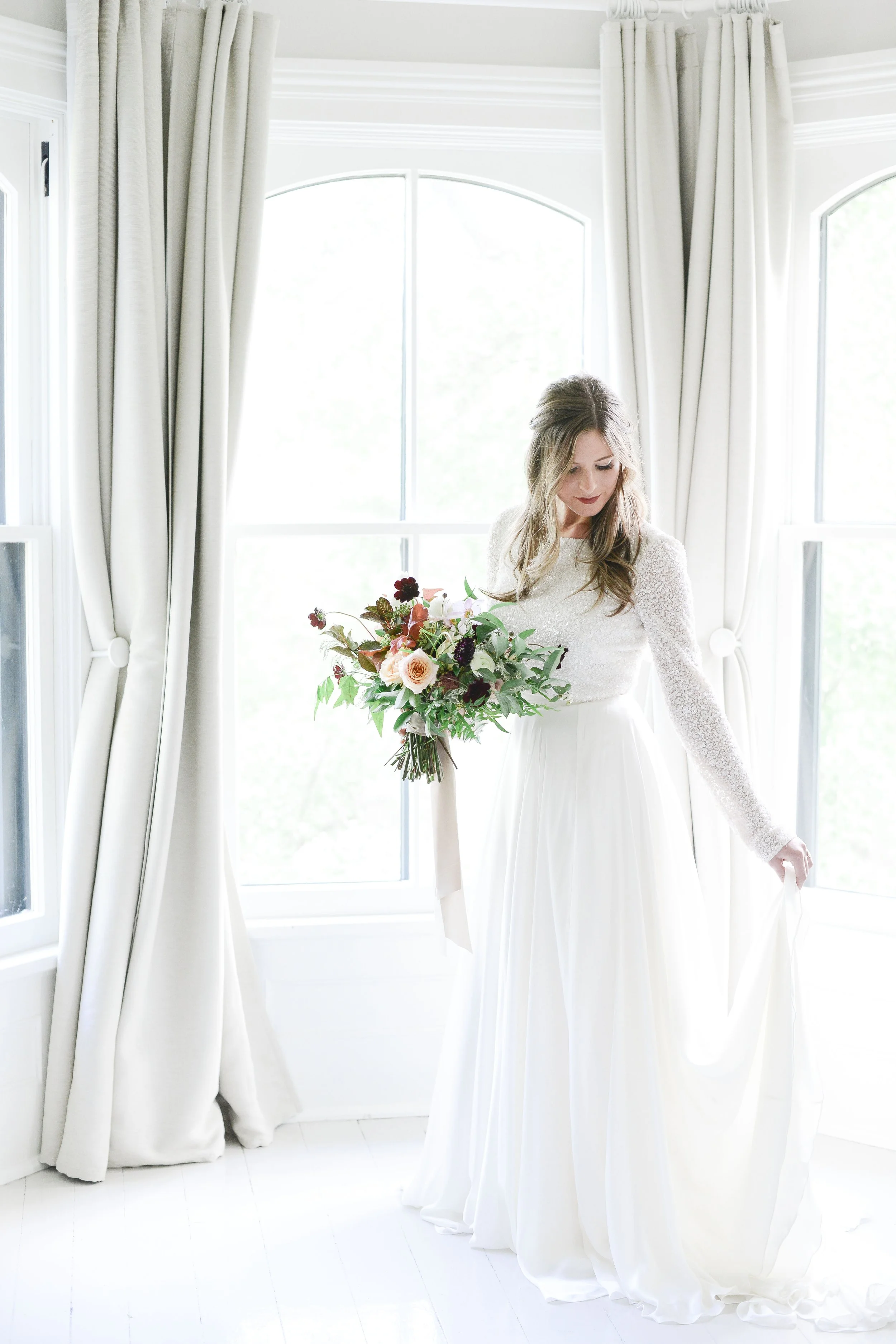 A woman with long hair wearing a white wedding gown holding a bouquet of flowers standing near large windows with white curtains in a bright room.
