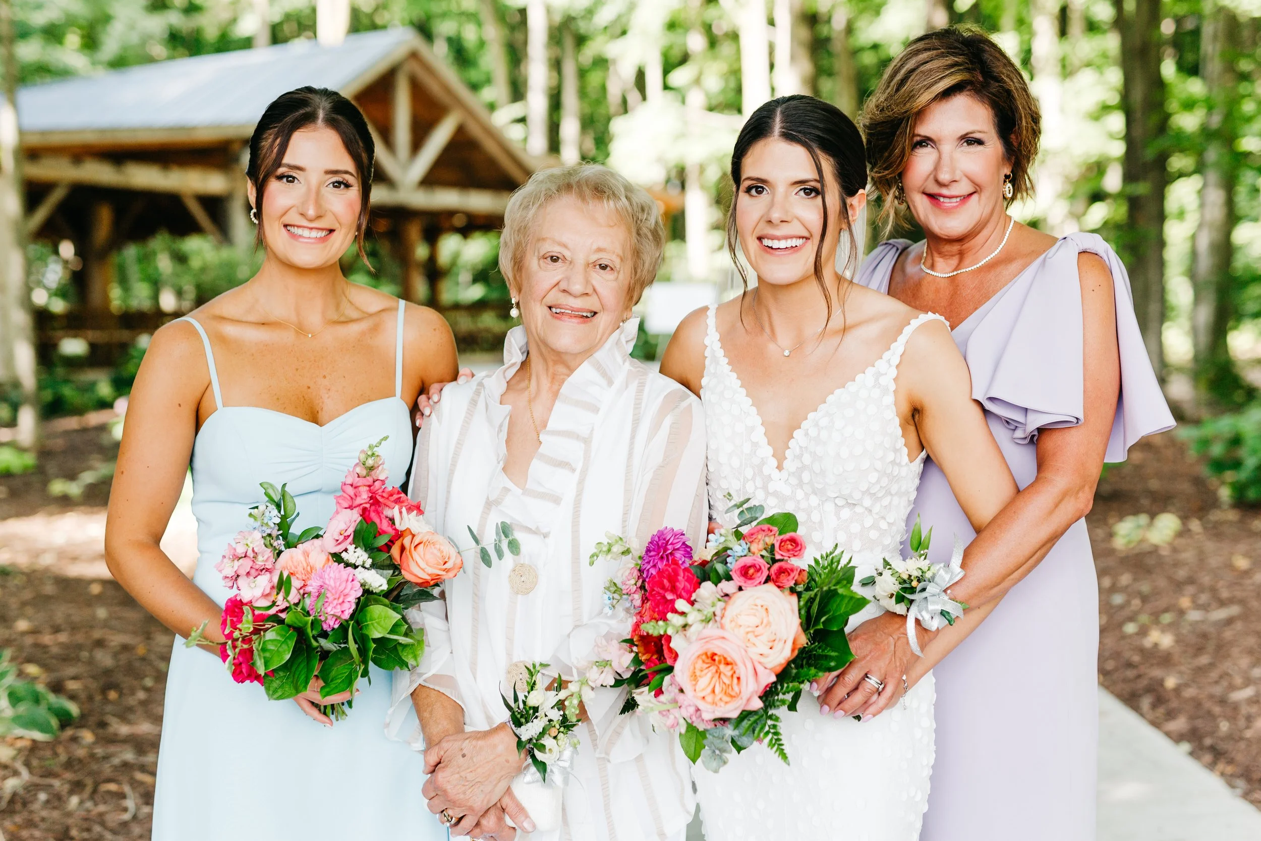 Group of four women, including a bride and family members, posing for a photo outdoors at Ivy Ridge Event Centre in the shaded wooded area, holding colourful bouquets of flowers.